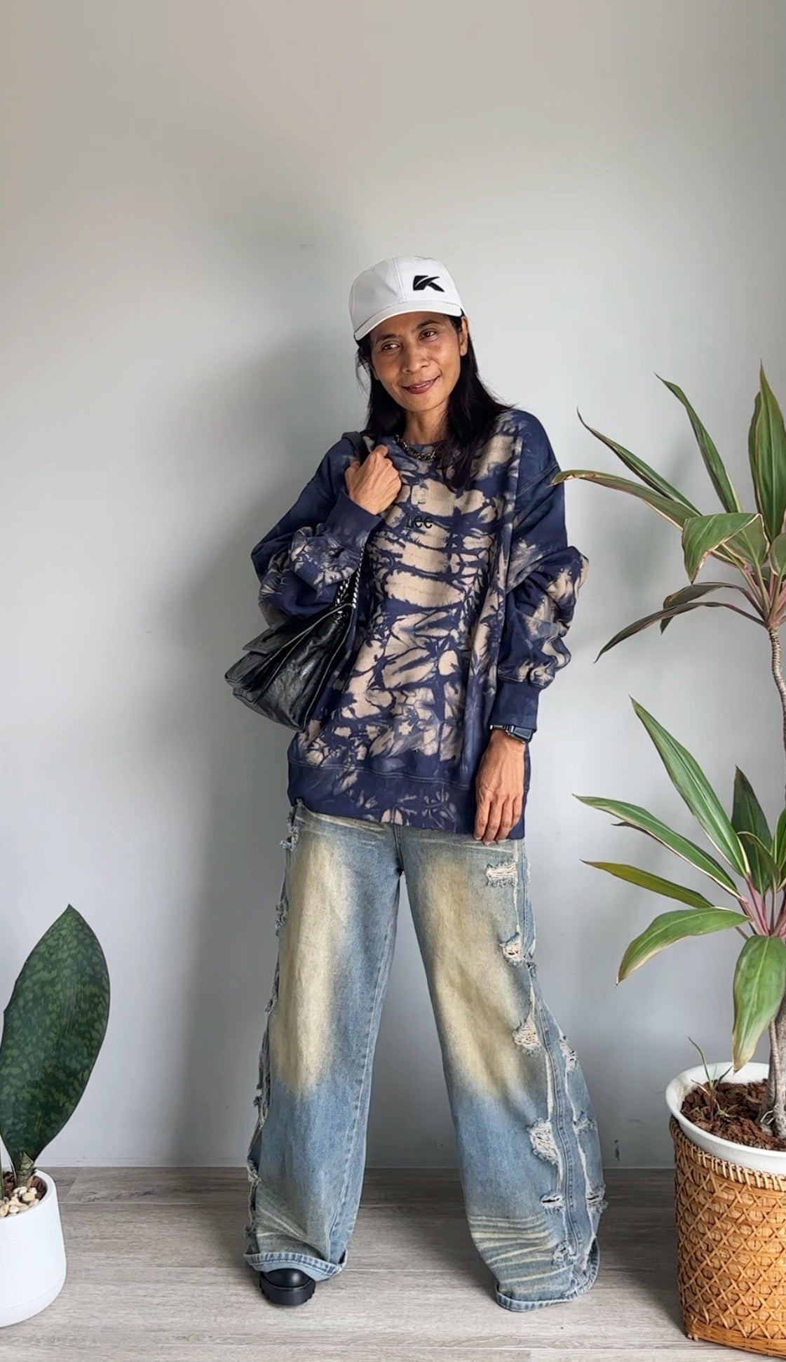 Person wearing a tie-dye sweatshirt and jeans standing indoors with plants around
