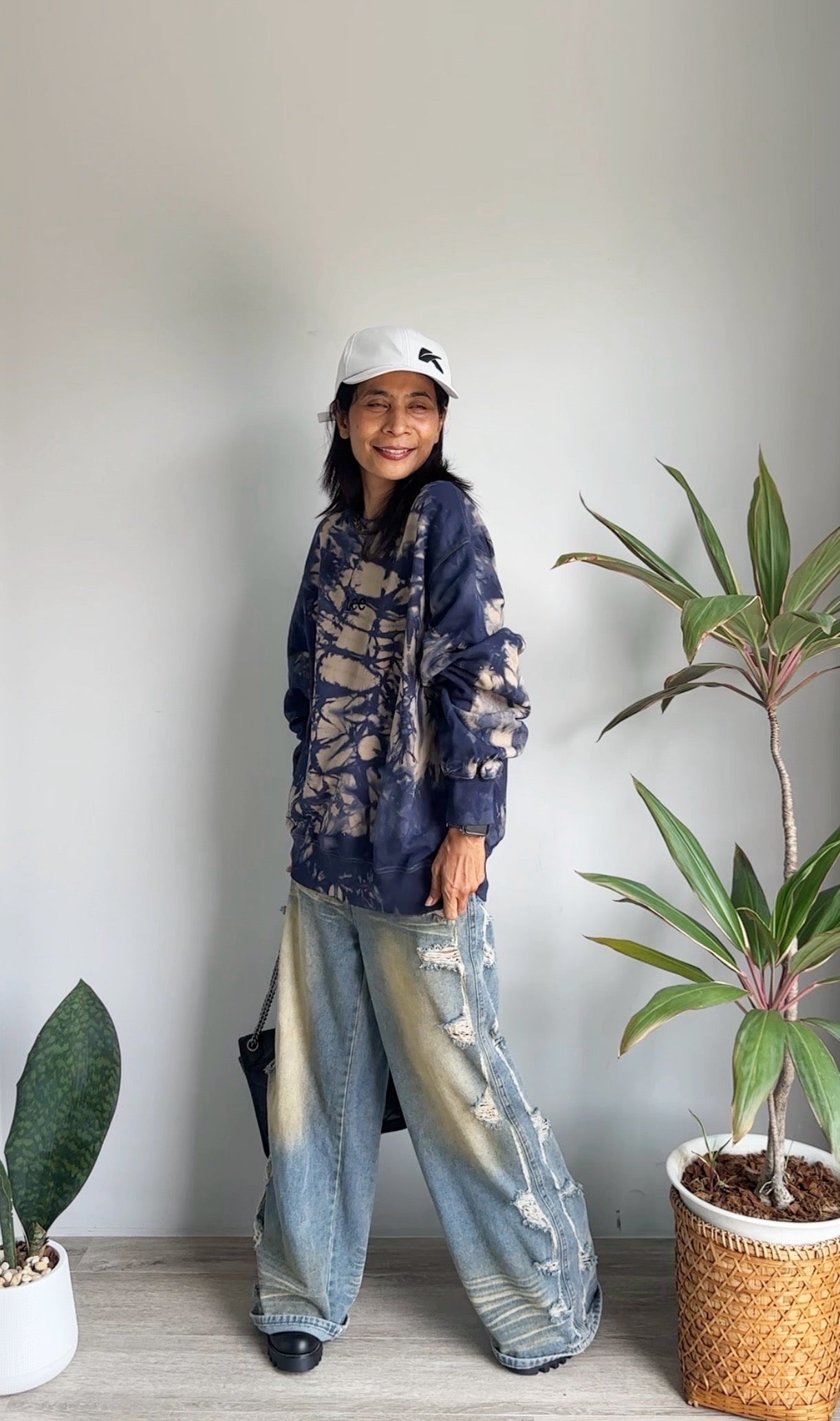 Person wearing a navy tie-dye sweater and light blue jeans standing indoors with plants around.