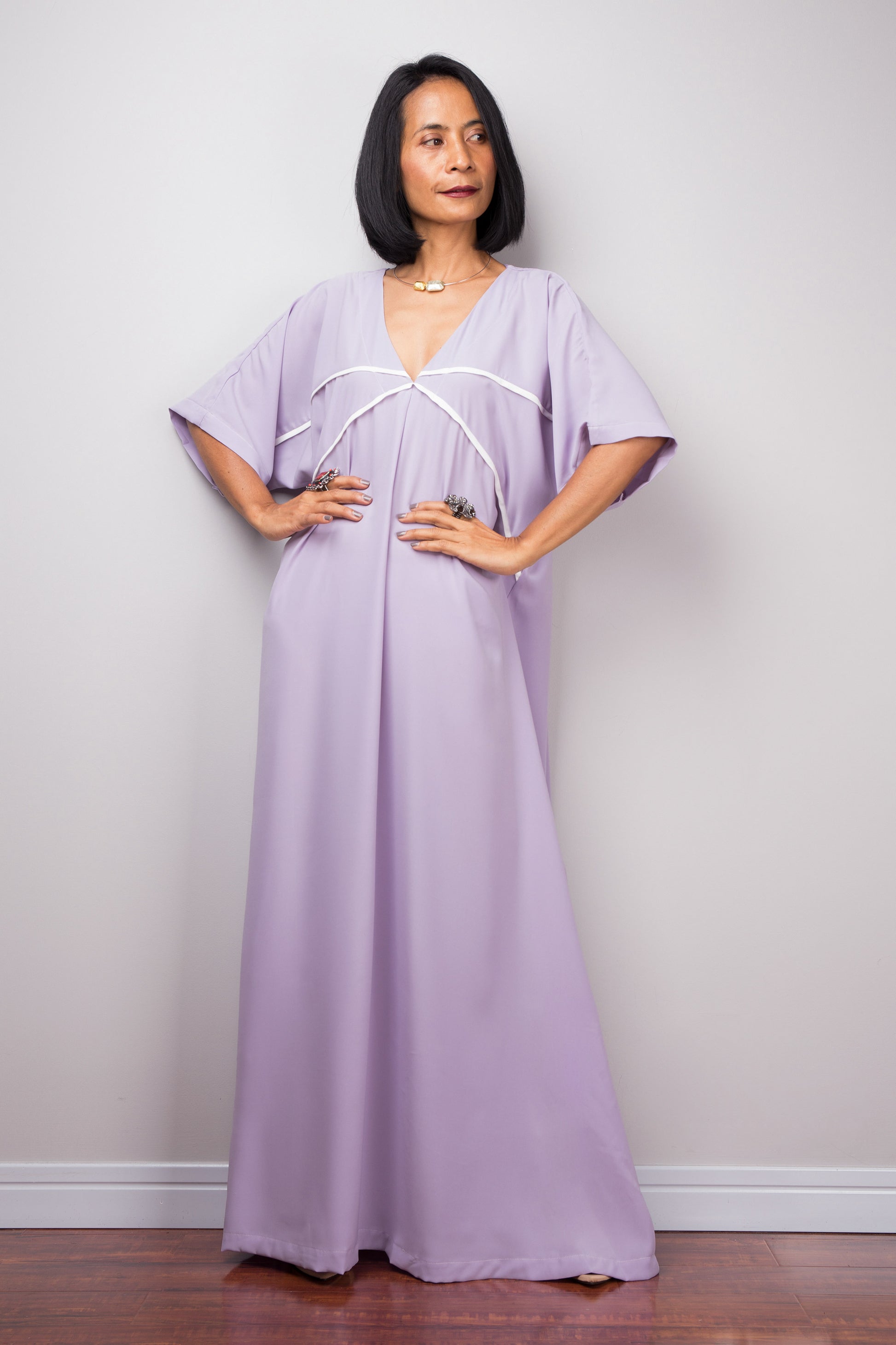 Woman wearing a lilac kaftan maxi dress with white crisscross trim, standing against a gray wall on a wooden floor, featuring short wide sleeves."


