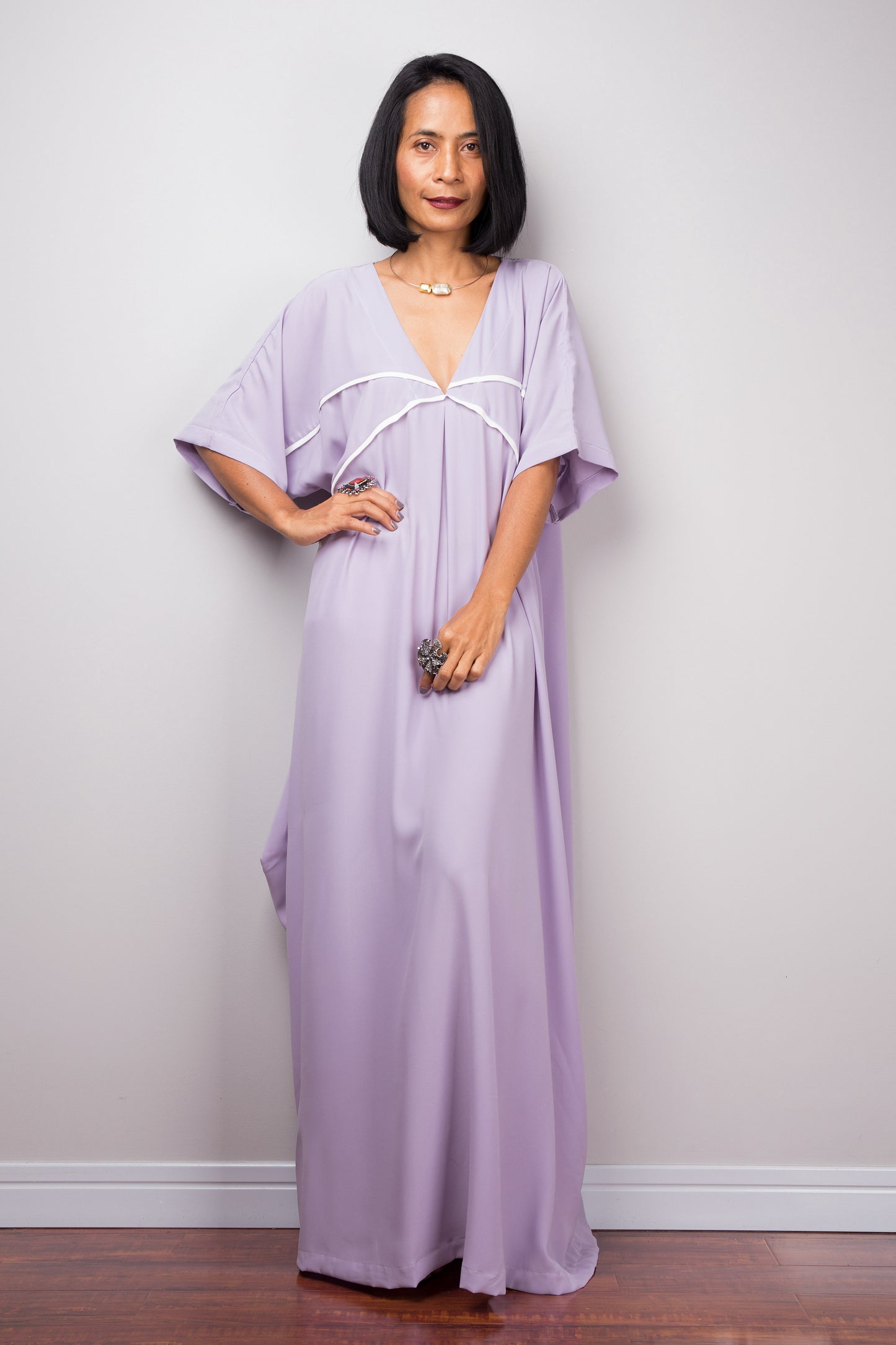 Nuichan’s lilac kaftan maxi dress with white trim, modeled by a woman with short black hair, posed in a minimalist studio, wearing heels, and a silver necklace.

