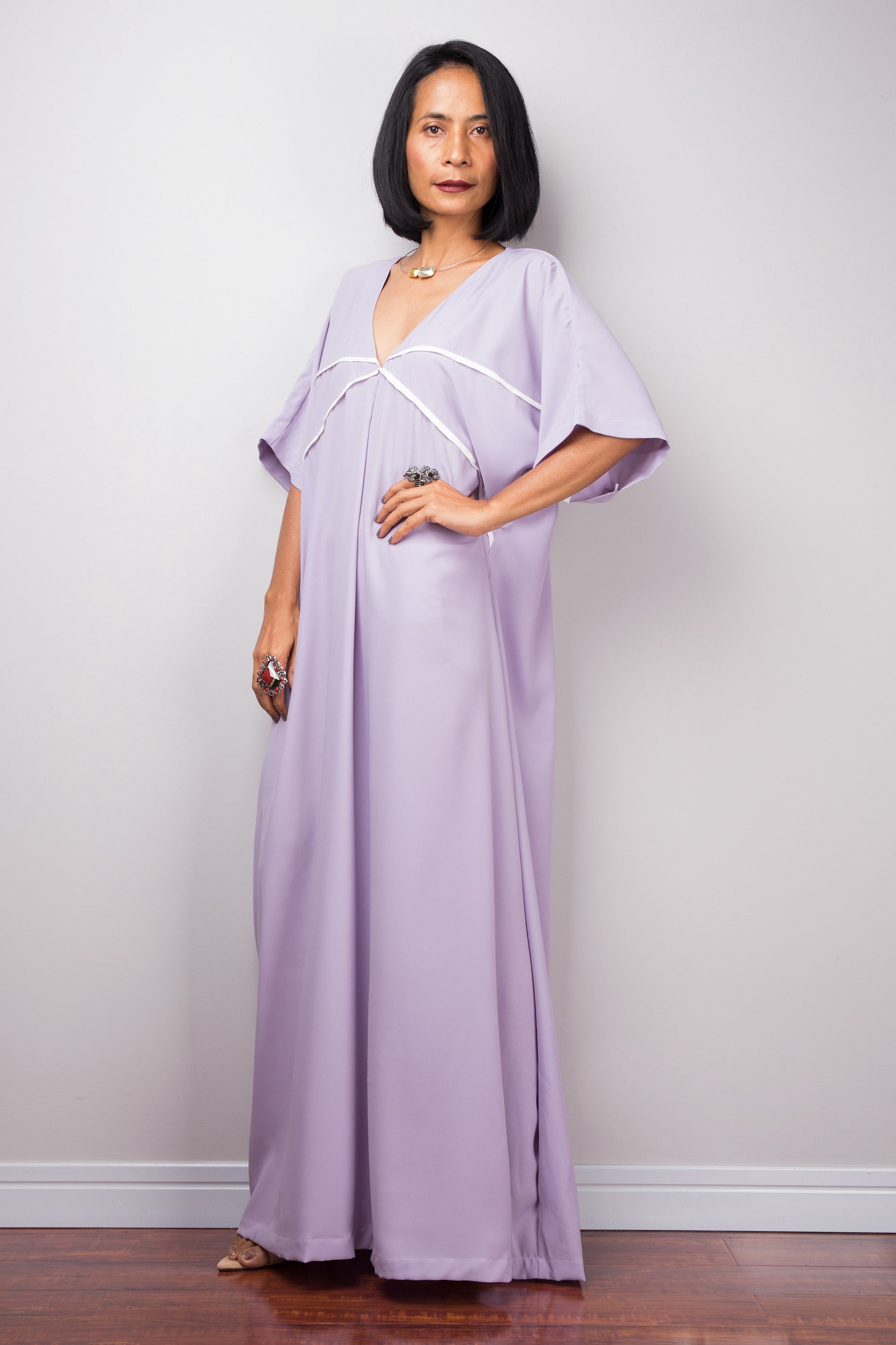A floor-length lilac kaftan dress from Nuichan, with white crisscross chest detail, worn by a woman standing sideways, set against a gray wall and wooden floor.

