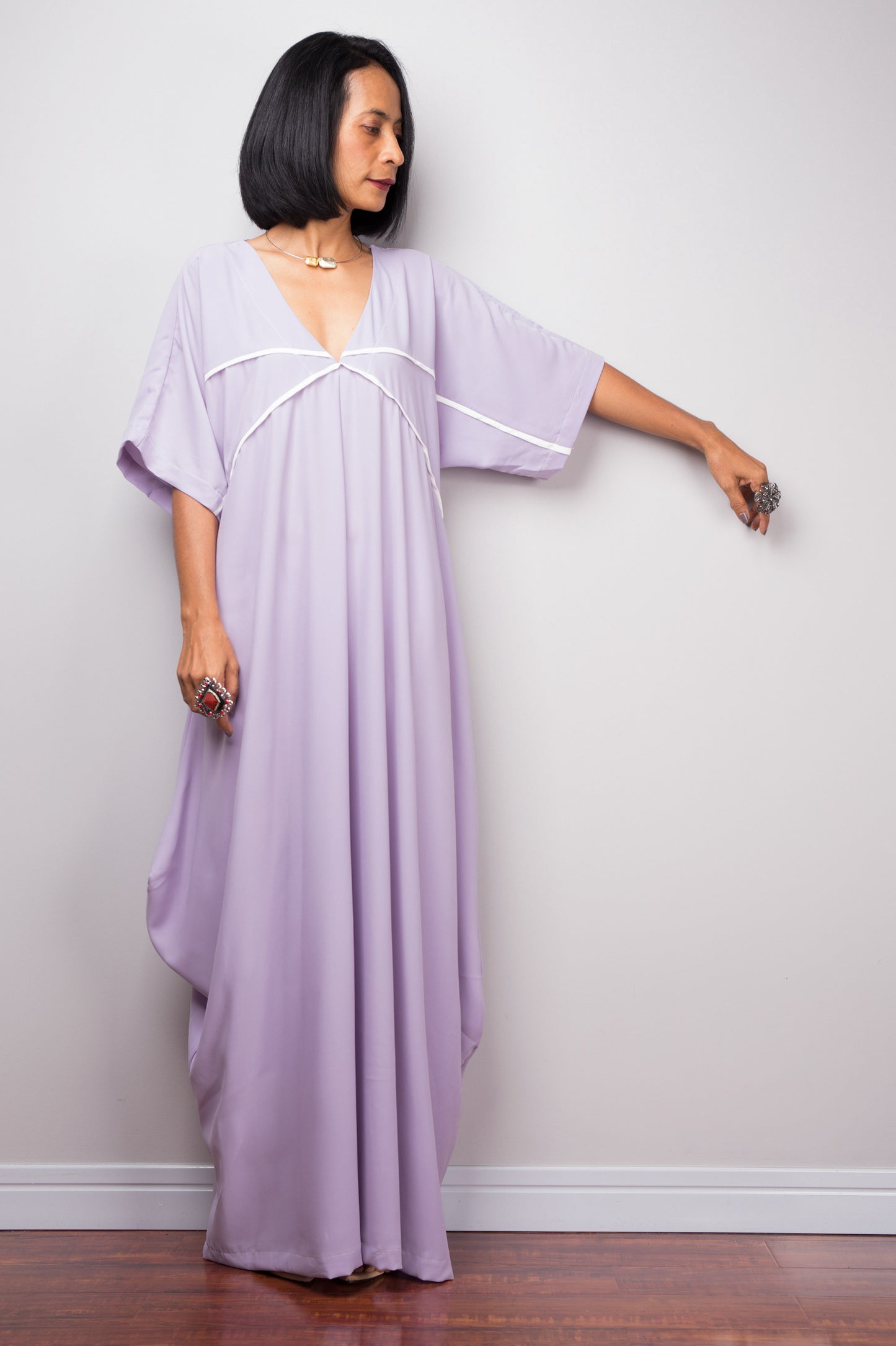 A woman modeling a lilac kaftan maxi dress from Nuichan, featuring a V-neck and wide sleeves with white trim, standing in a simple gray studio

