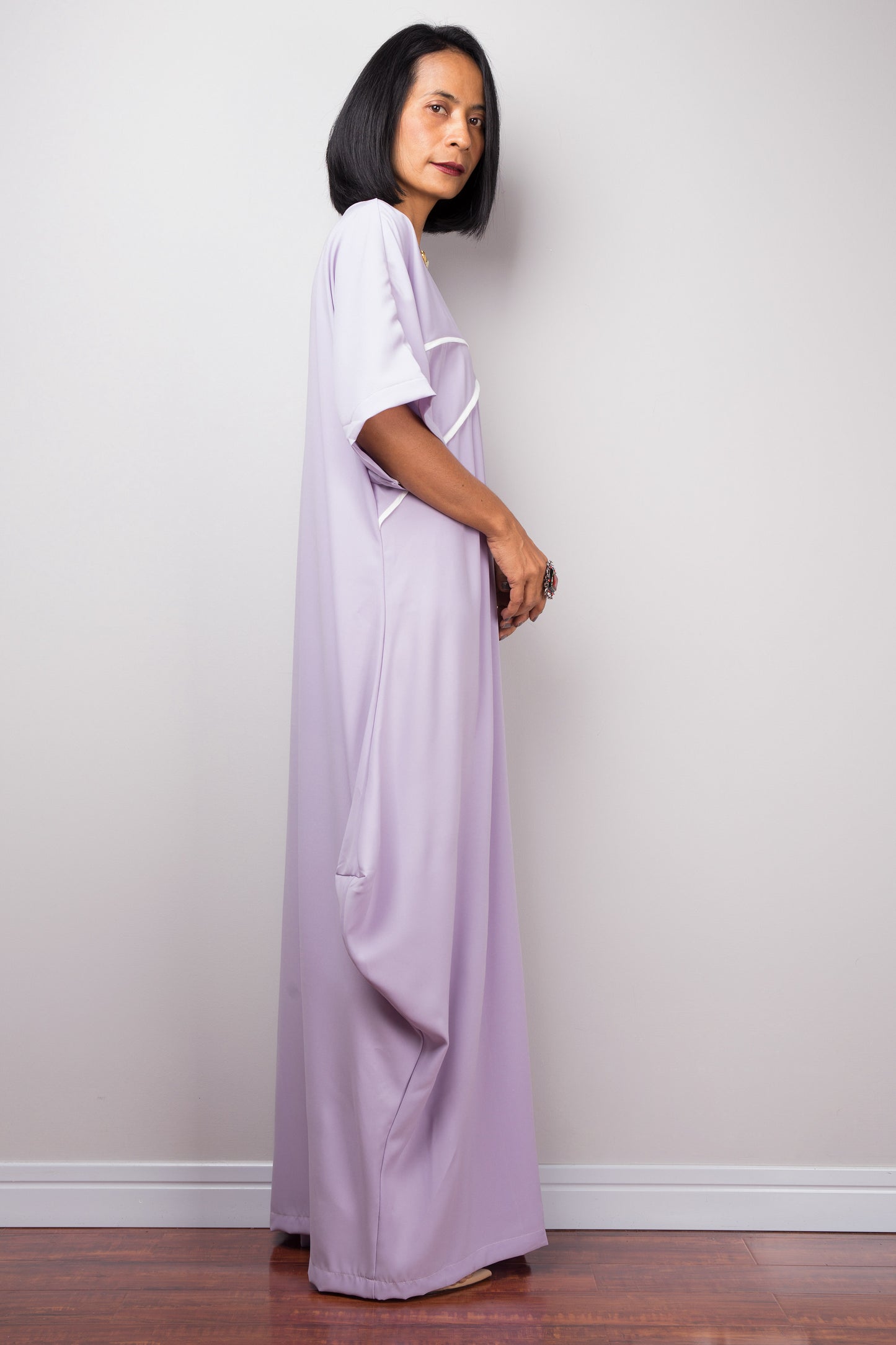 A floor-length lilac kaftan dress from Nuichan, with white crisscross chest detail, worn by a woman standing sideways, set against a grey wall and wooden floor.

