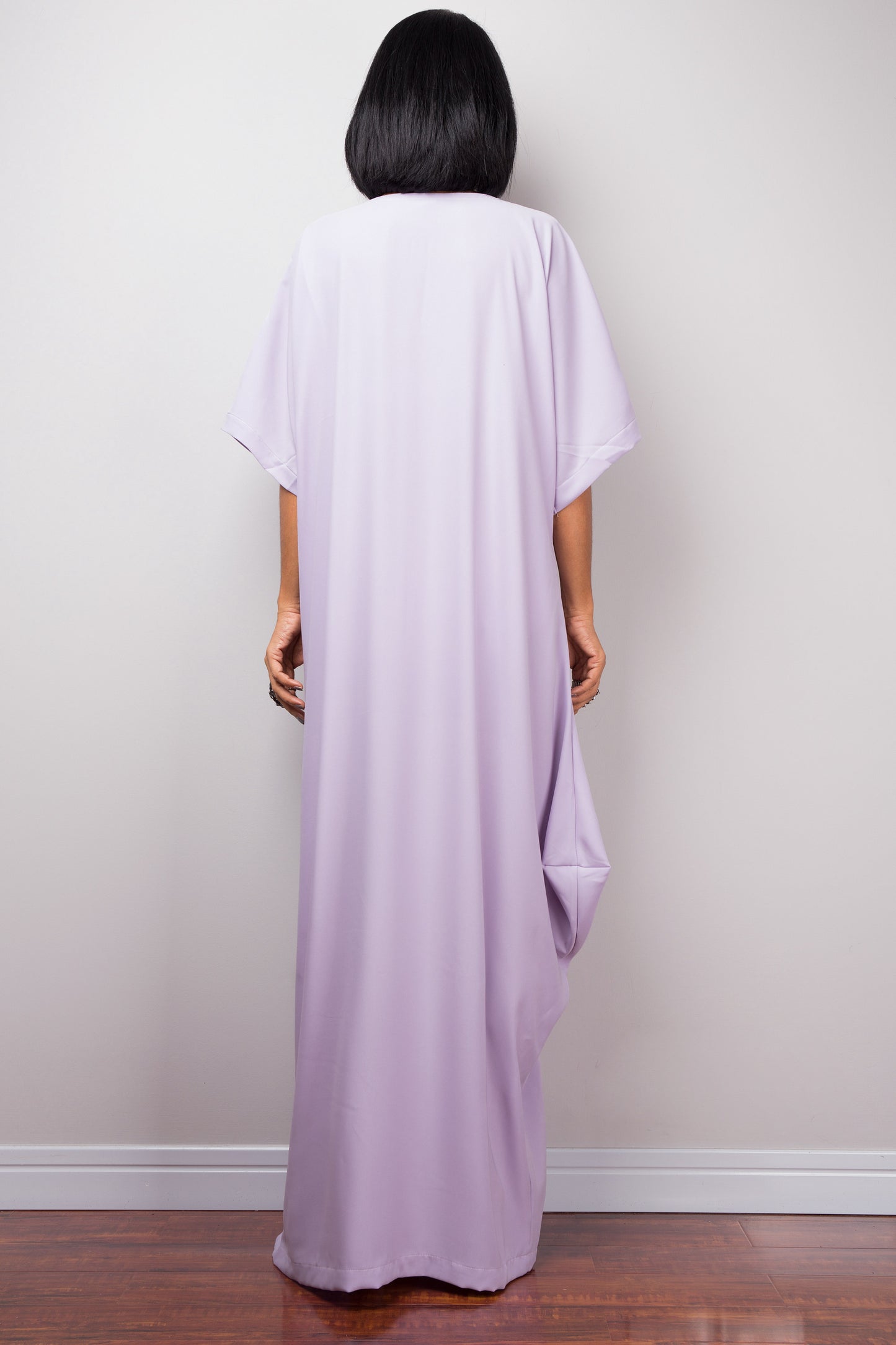 Nuichan’s floor-length lilac kaftan dress with white crisscross detail, modeled by a woman in a studio with a gray background and wooden floor, back view

