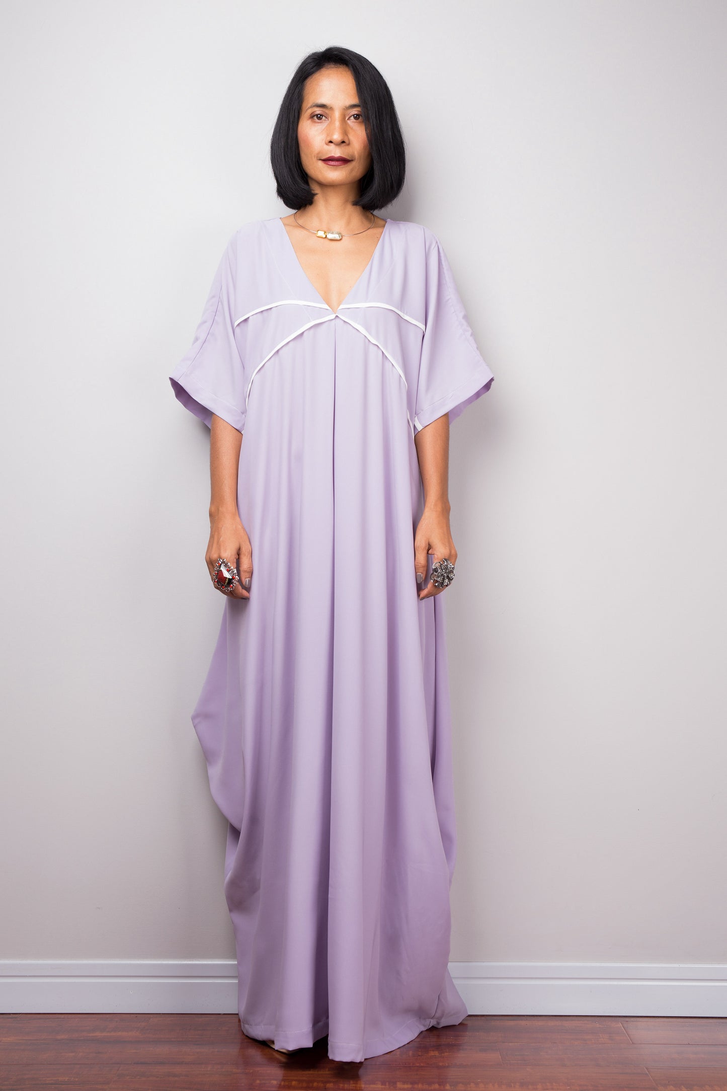 Nuichan’s floor-length lilac kaftan dress with white crisscross detail, modeled by a woman in a studio with a gray background and wooden floor

