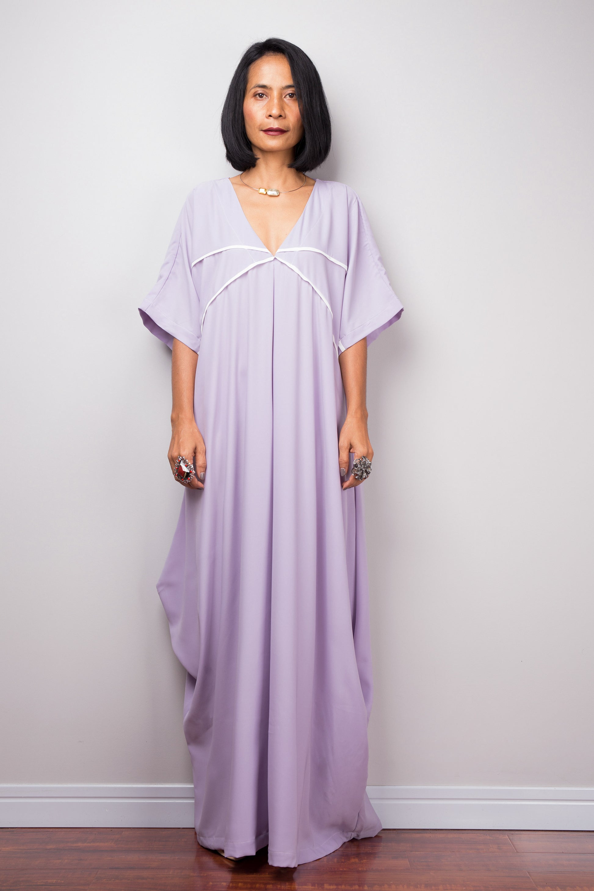 Nuichan’s floor-length lilac kaftan dress with white crisscross detail, modeled by a woman in a studio with a gray background and wooden floor

