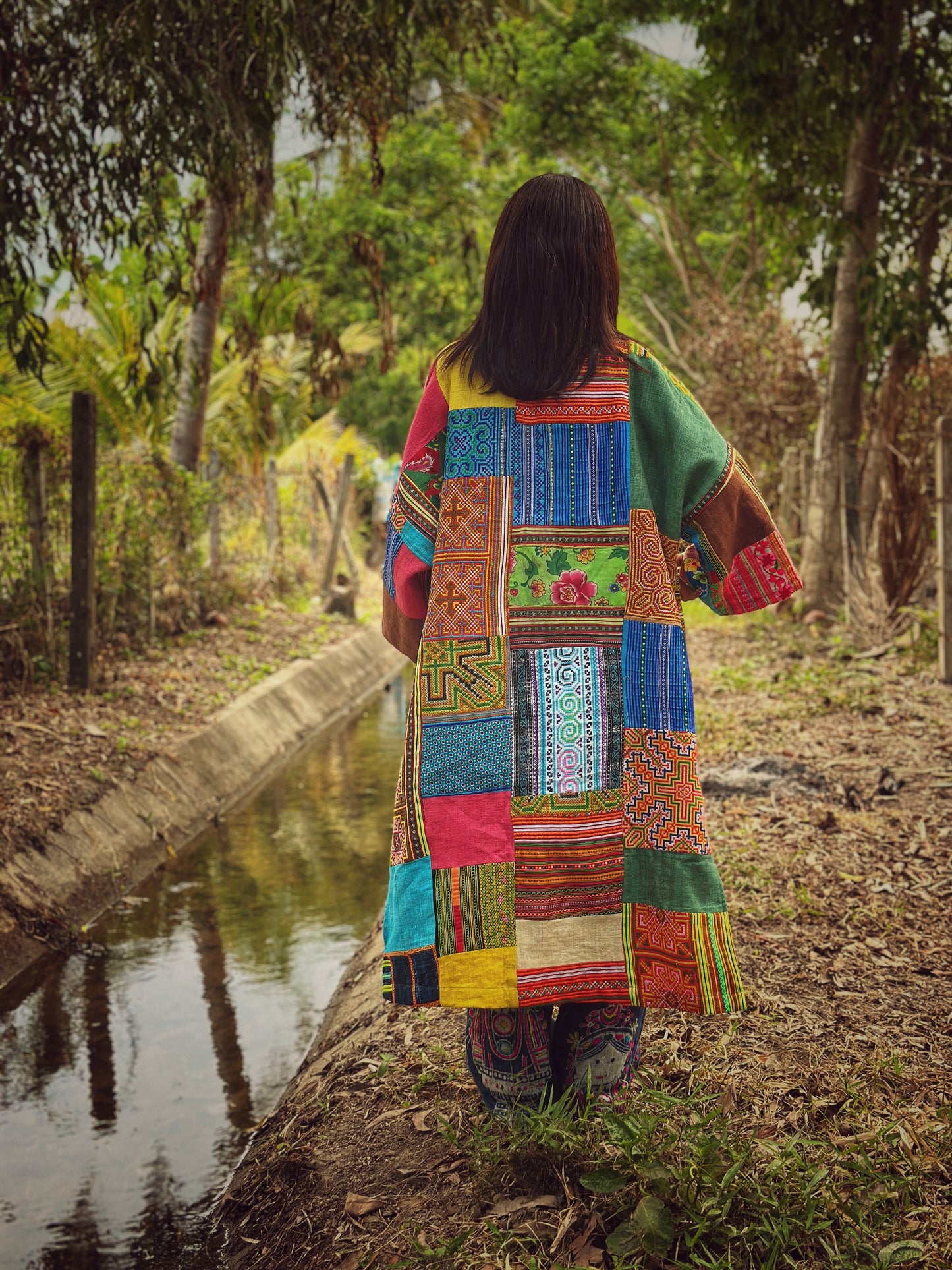 Woman wearing an open-front patchwork coat from hill tribe fabrics, adorned with intricate ethnic patterns in blue, red, and yellow. This boho coat is ideal for festivals, casual outings, or travel, photographed in a natural forest setting near a peaceful stream

