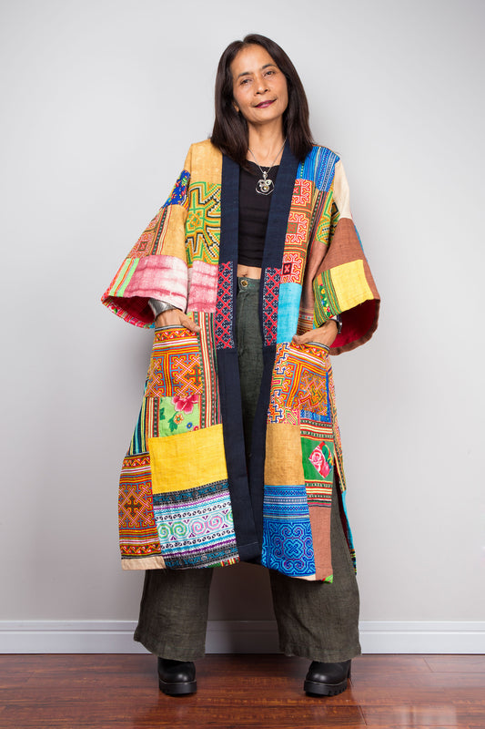 Nuichan wearing an open-front patchwork coat made from colorful hill tribe fabrics, featuring intricate ethnic patterns in blue, red, and yellow hues. 