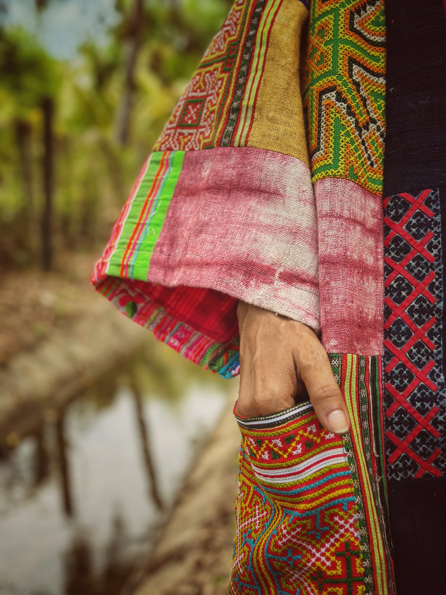 Close-up of an open-front patchwork coat made from hill tribe fabrics, showcasing detailed handwoven patterns in green, pink, and blue with floral and geometric designs. This artisanal boho coat, worn by a model in a forest, reflects the rich cultural heritage of hill tribe textiles.

