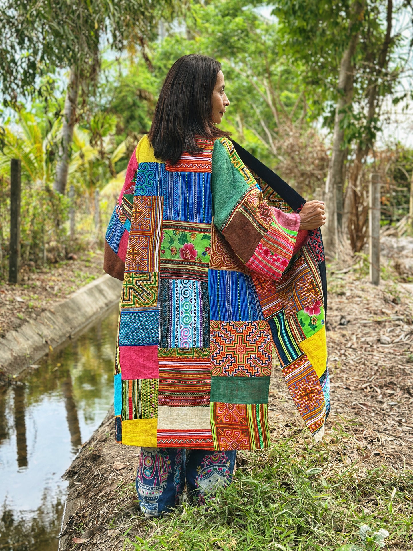 Back view of a woman in an open-front patchwork coat crafted from sustainable hill tribe fabrics, showcasing a vibrant mix of handwoven textiles in green, pink, and blue. This eco-friendly boho coat by Nuichan, made from upcycled materials, stands out against a natural forest backdrop near a stream


