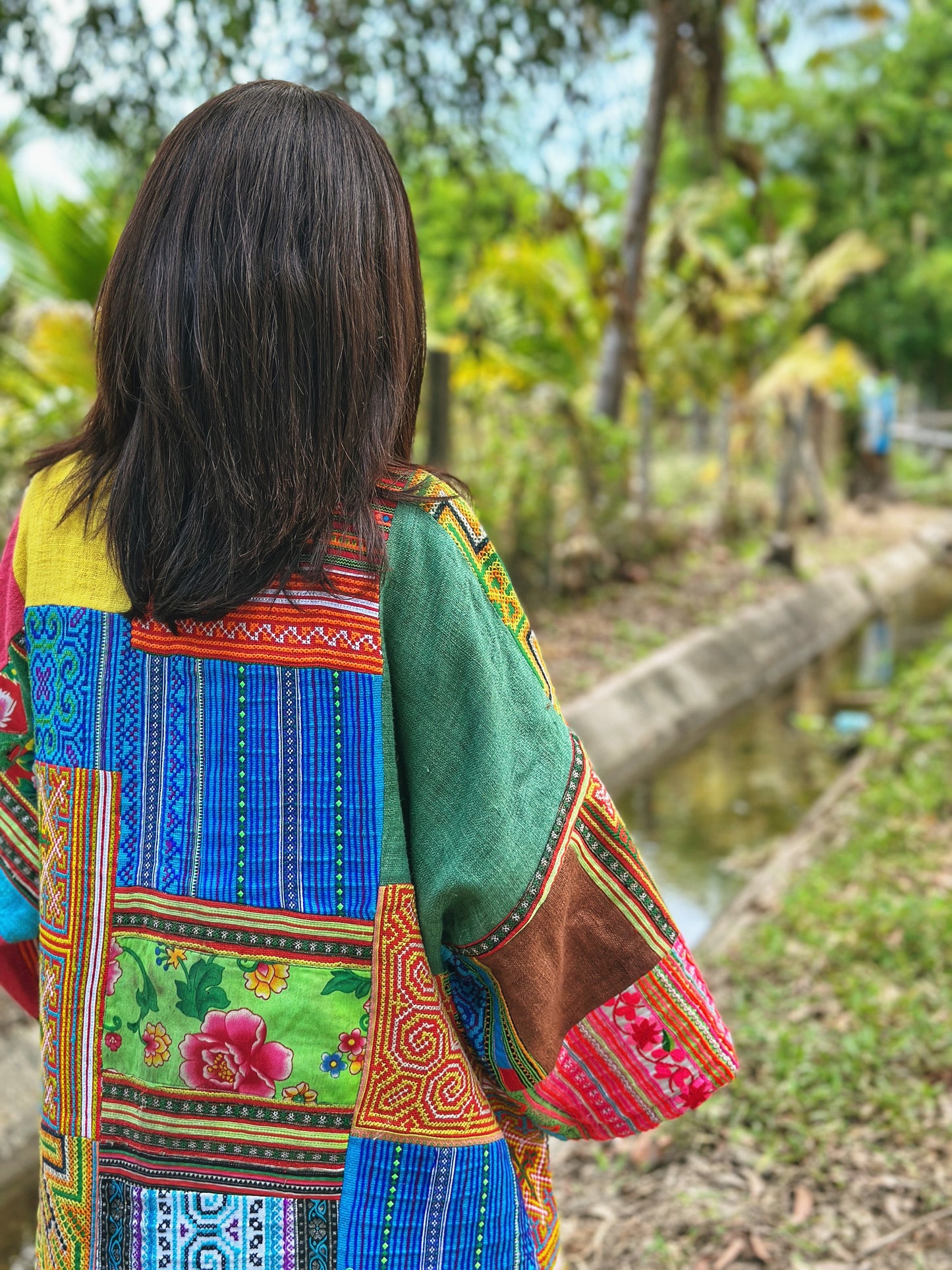 Woman in an open-front patchwork coat made from hill tribe fabrics, featuring a colorful mix of tribal patterns in green, blue, and red. This boho coat, perfect for spring or fall, is captured in a lush forest setting near a stream, highlighting its earthy, natural appea

