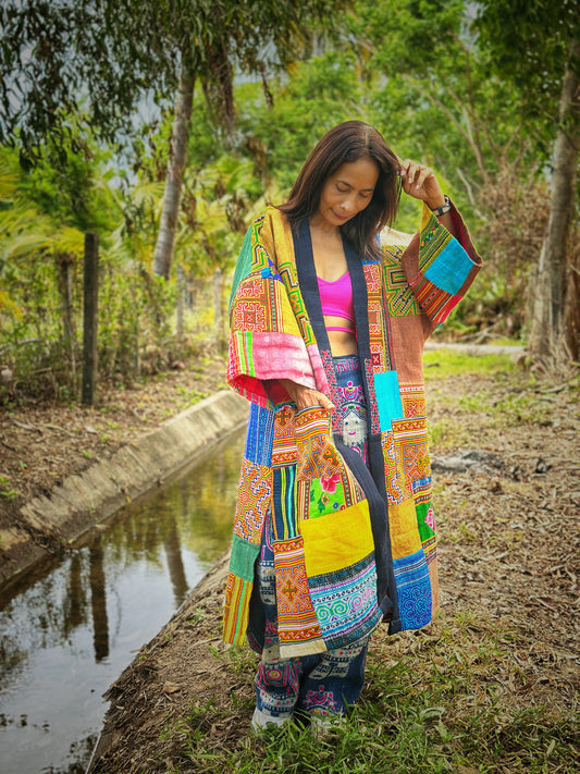 Model wearing an open-front patchwork coat made from colorful hill tribe fabrics, featuring intricate ethnic patterns in blue, red, and yellow hues. This boho coat by Nuichan, with its vibrant geometric and floral designs, is perfect for adding a unique, artisanal touch to any outfit in a lush forest setting


