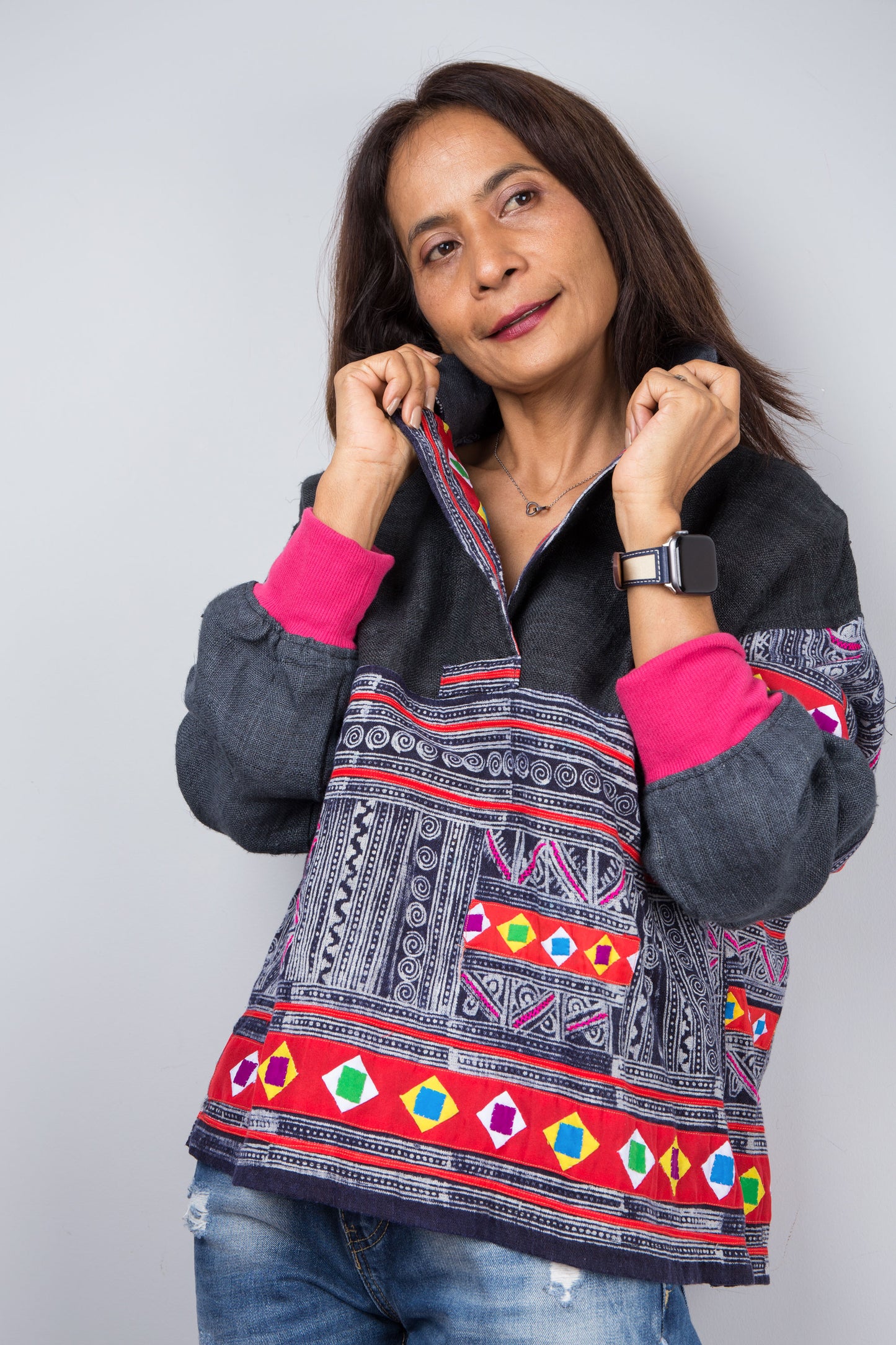  Hand-batik pullover tunic with colorful patchwork and pink cuffs on a woman, paired with distressed jeans, set against a gray wall and wooden floor, by Nuichan.

