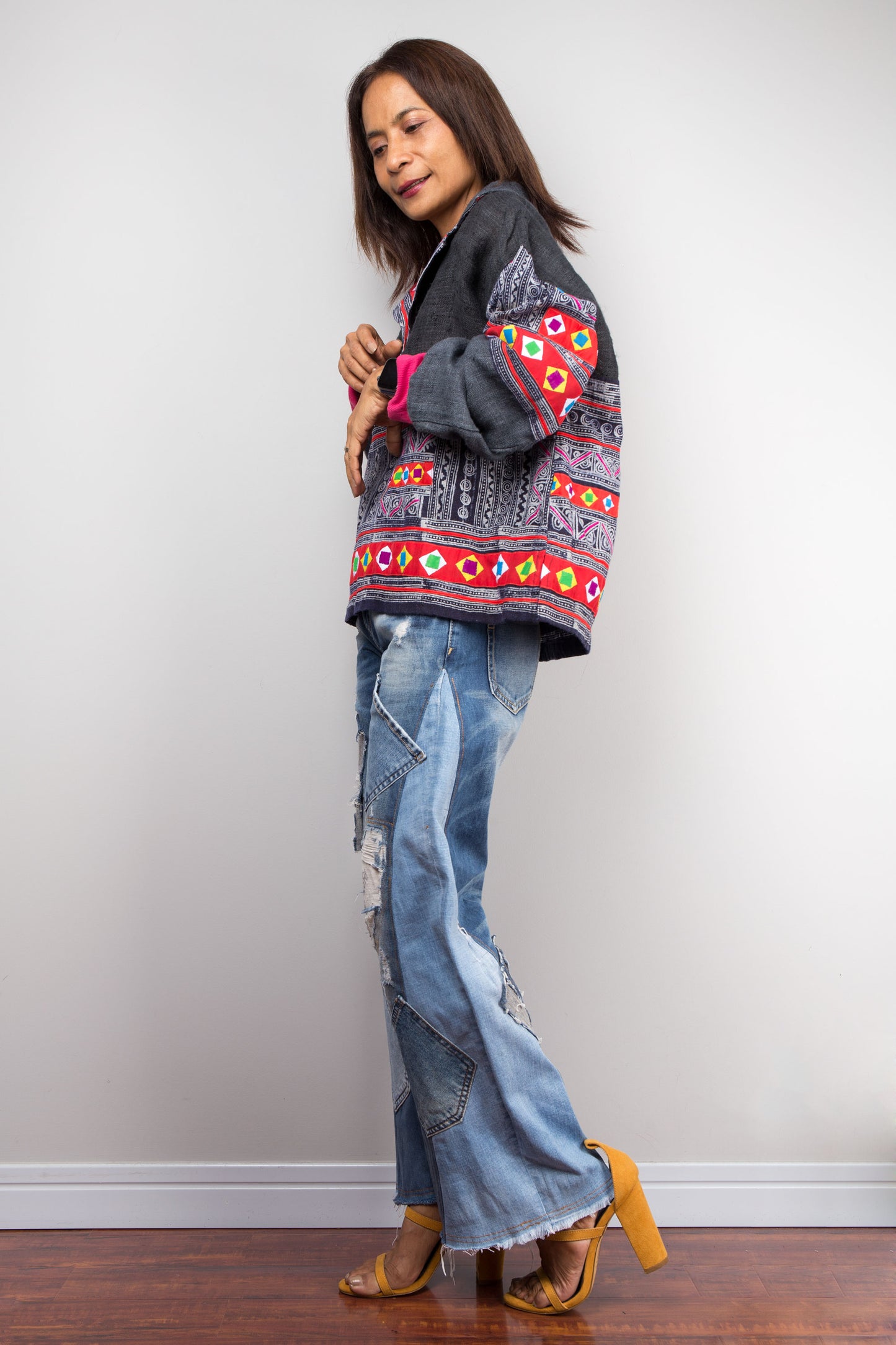 Model in a hand-batik pullover tunic with pink cuffs and patchwork by Nuichan, styled with jeans, against a gray wall and wooden floor, accessorized with tan heeled sandals.

