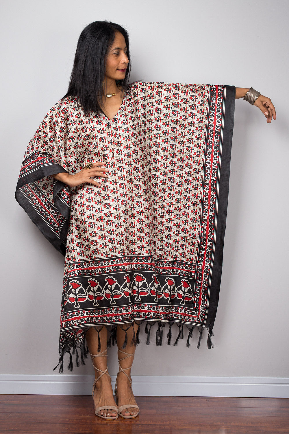 Woman wearing a petite caftan dress with pattern and fringe against a white wall