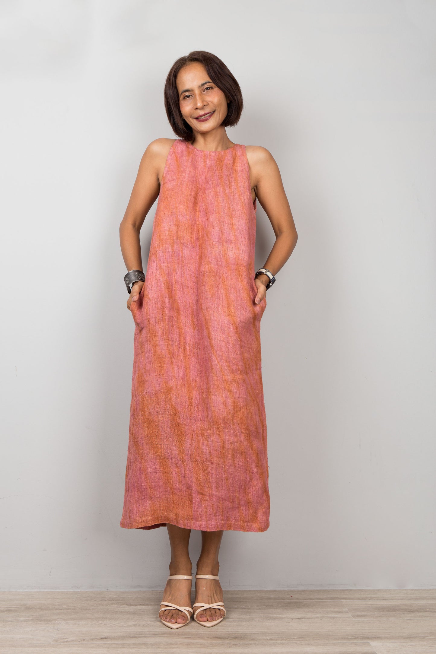 Woman wearing a pink coral dress standing against a plain background.  Sleeveless hemp dress with shibori tie dye details.