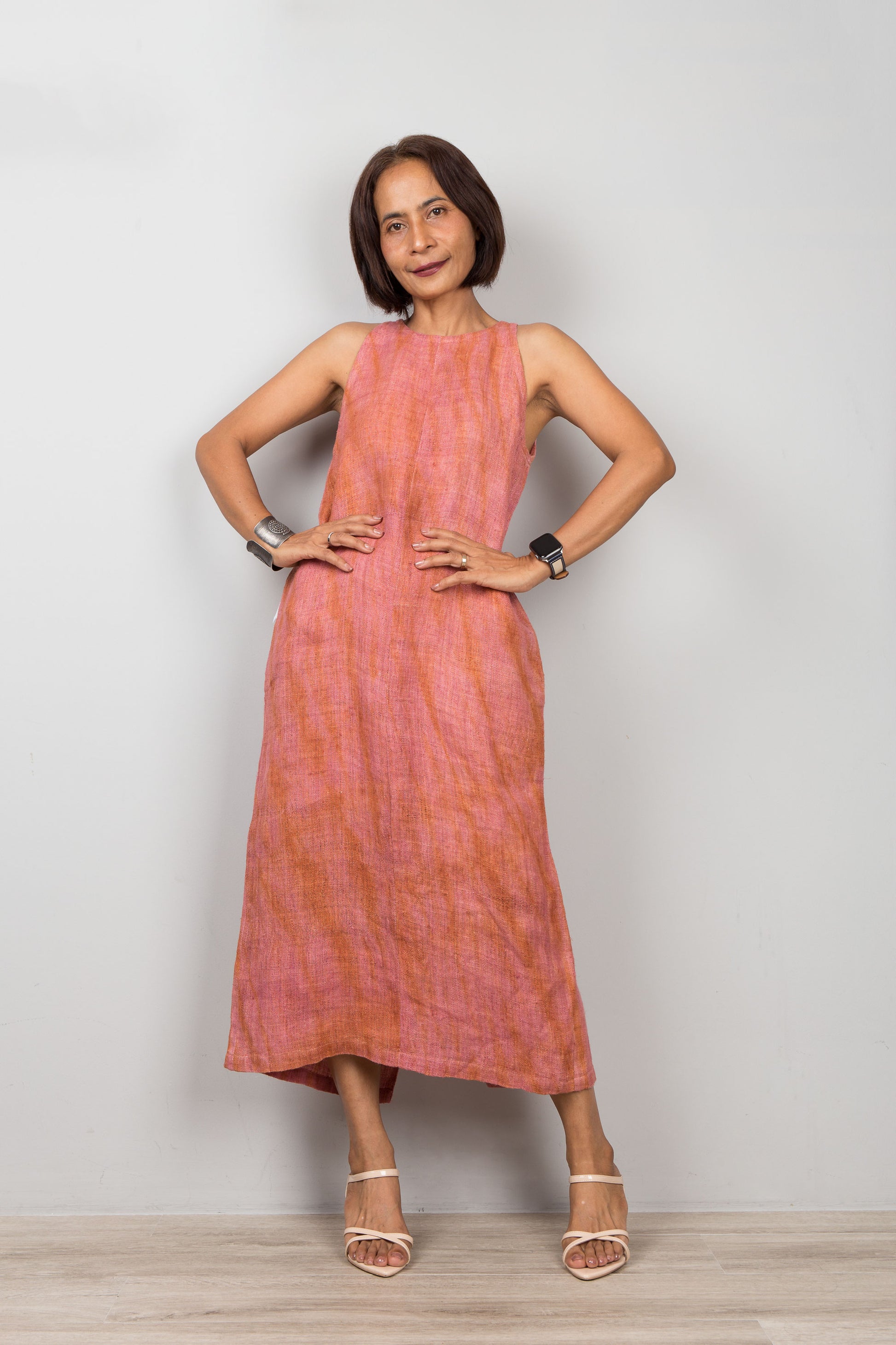 Woman wearing a sleeveless pink coral dress standing against a plain background.  Organic hemp dress with shibori tie dye details.