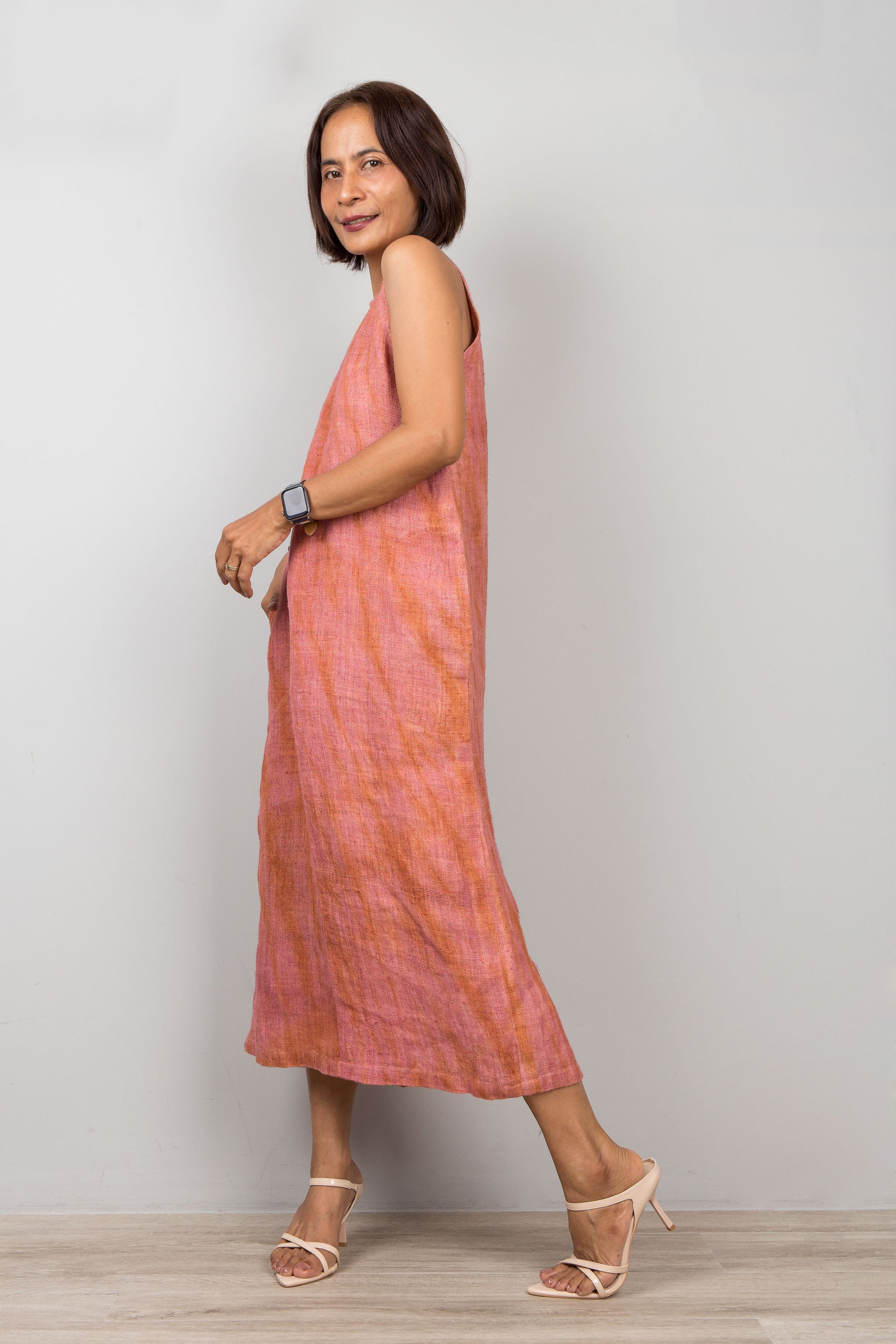 Woman wearing a soft coral dress standing against a plain background.  Sleeveless hemp dress with shibori tie dye details.