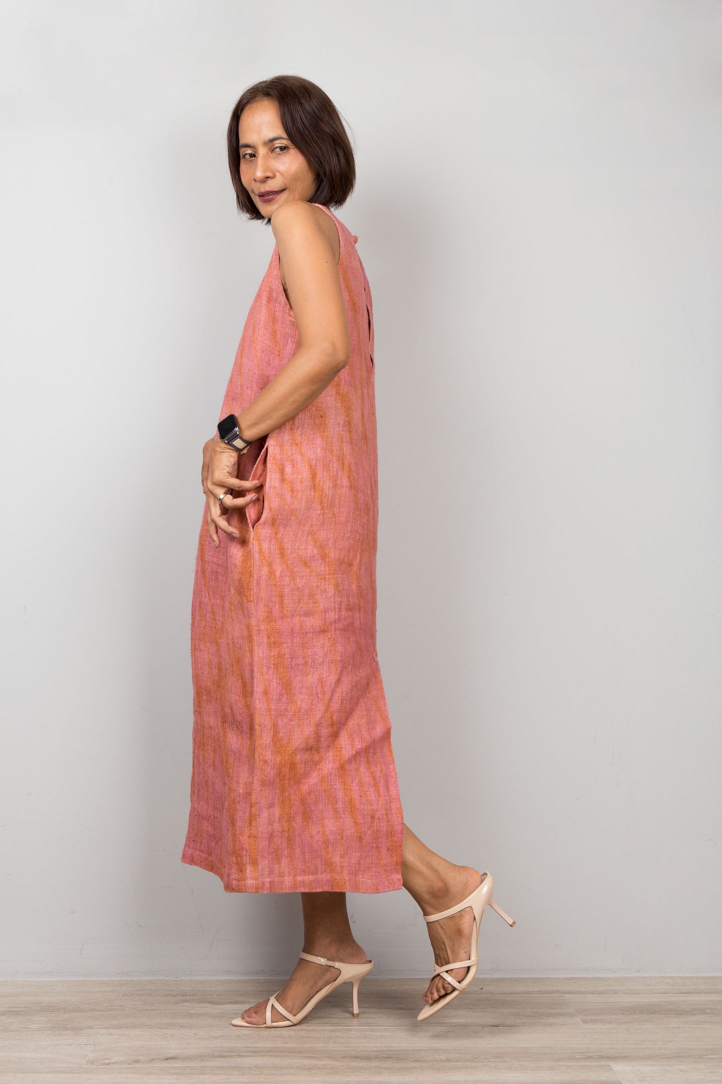 Woman wearing a sleeveless pink coral dress standing against a plain background.  Handwoven hemp dress with shibori tie dye details.