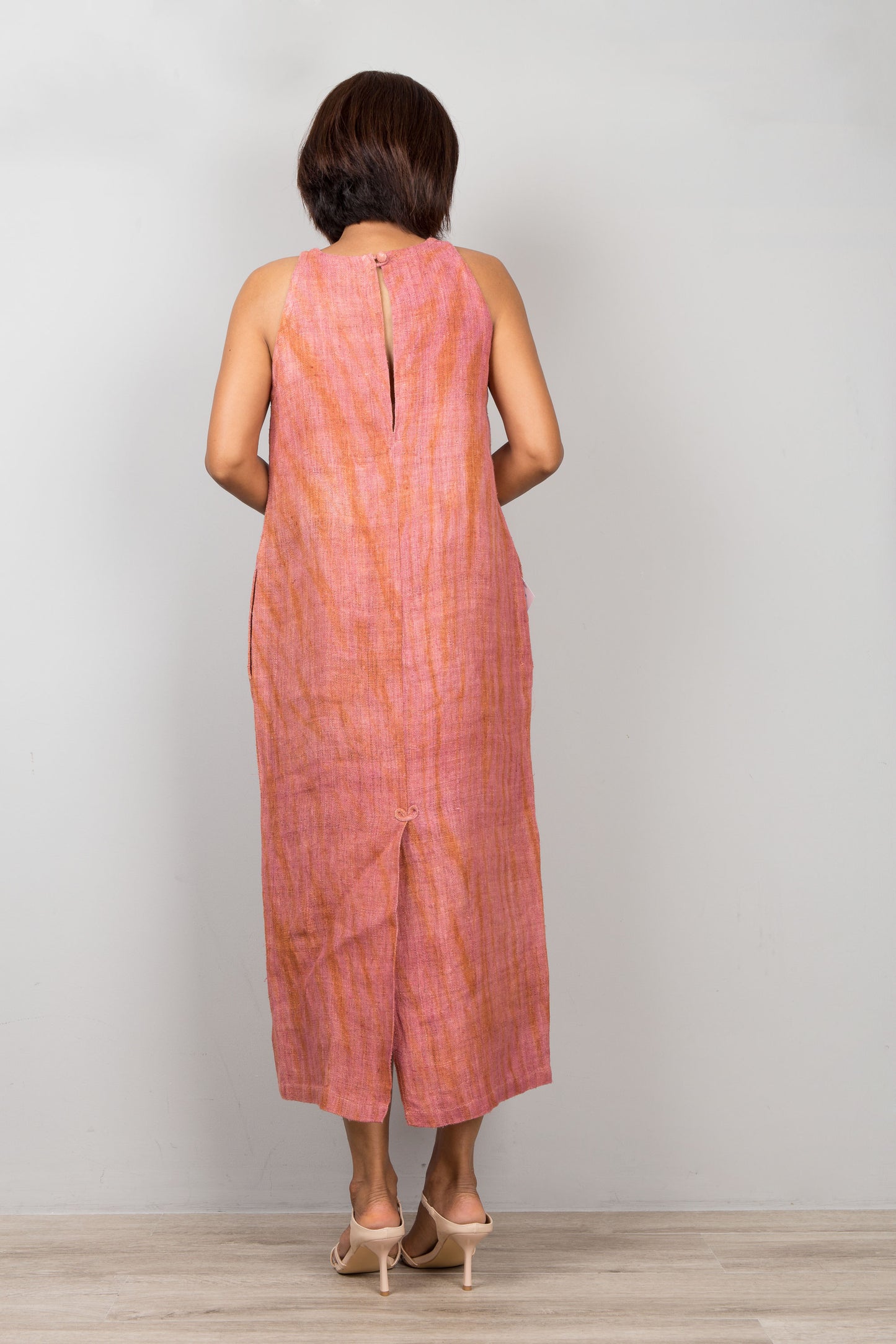 Woman wearing a sleeveless coral dress standing against a plain background.  Handwoven hemp dress with shibori tie dye details.