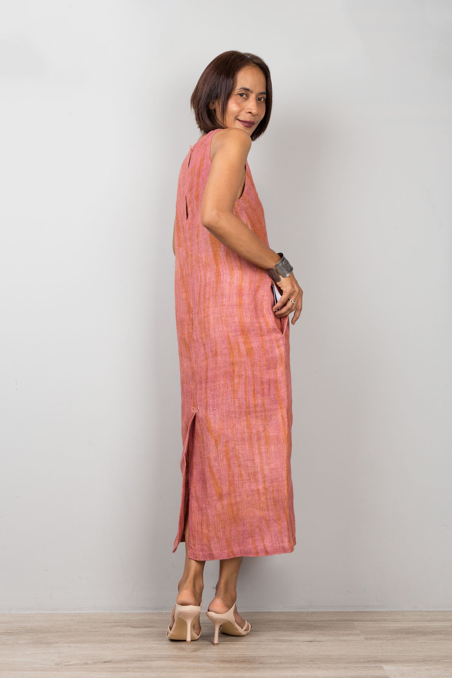 Woman wearing a sleeveless pink coral dress standing against a plain background.  Handwoven hemp midi dress with shibori tie dye details.