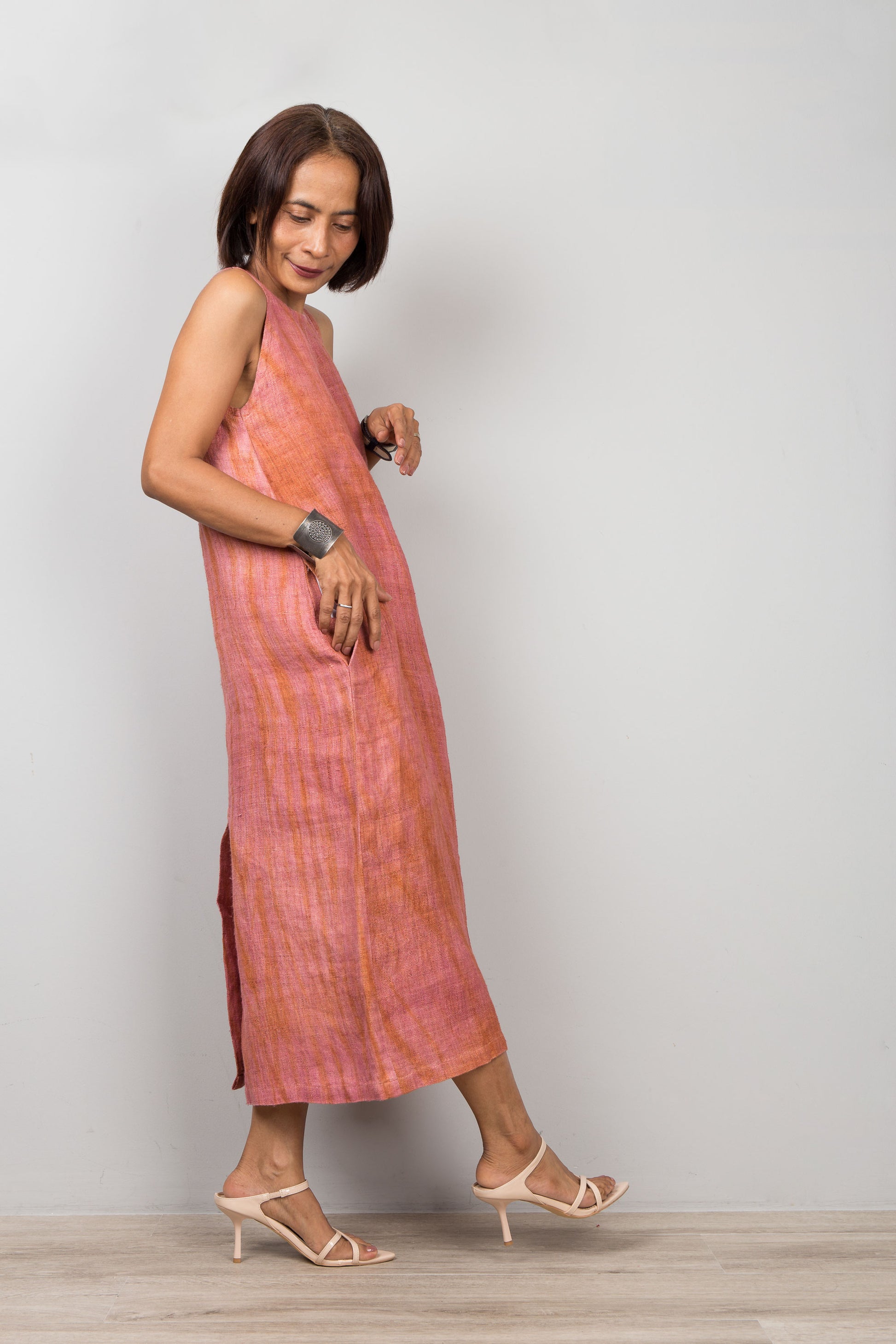 Woman wearing a sleeveless pink coral dress for petite standing against a plain background.  Handwoven hemp dress with shibori tie dye details.