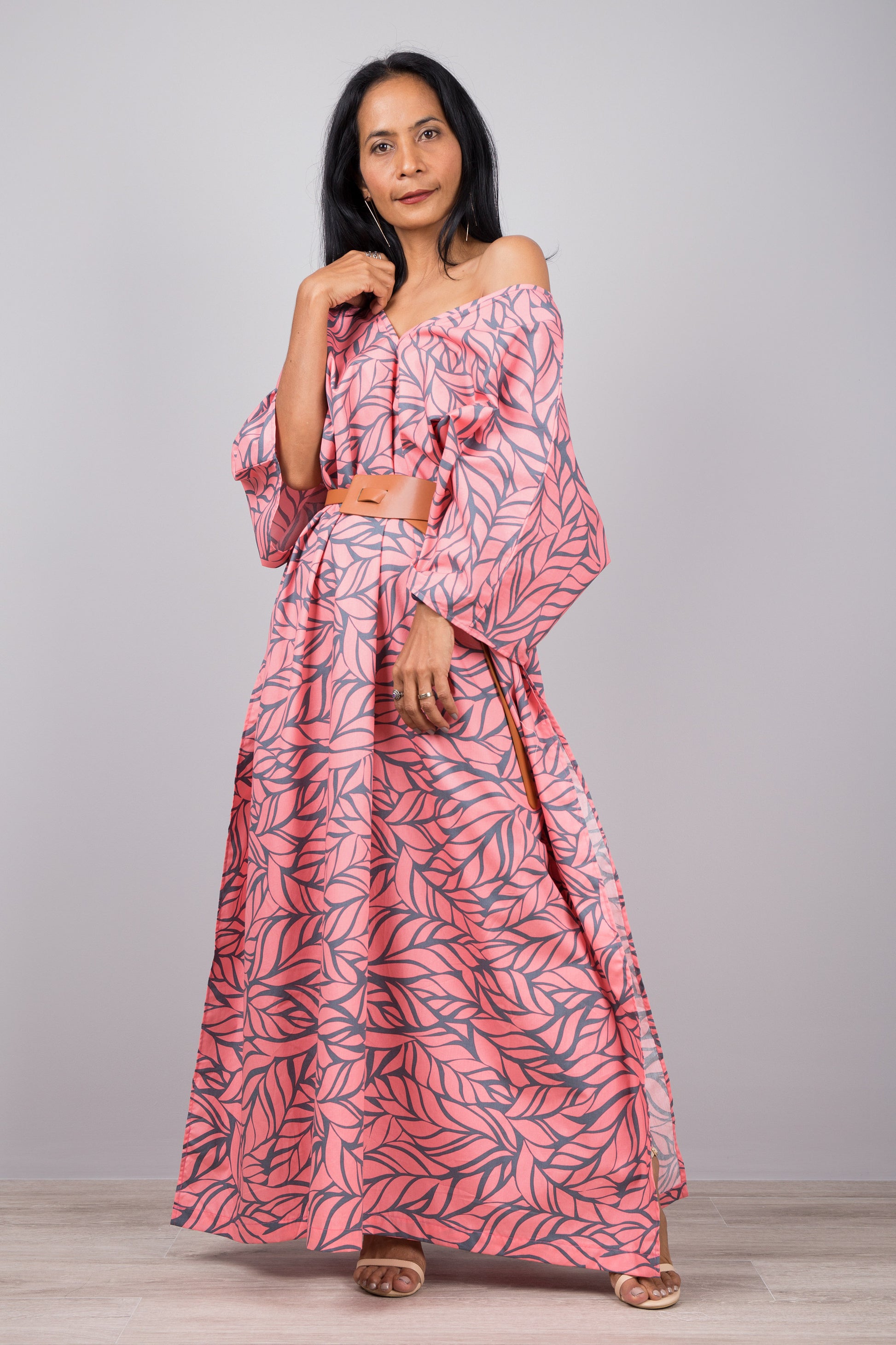 Woman in a tropical pink kaftan dress with gray leaf design and side split, posed against a gray wall with a wooden floor, wearing a smartwatch and delicate necklace.

