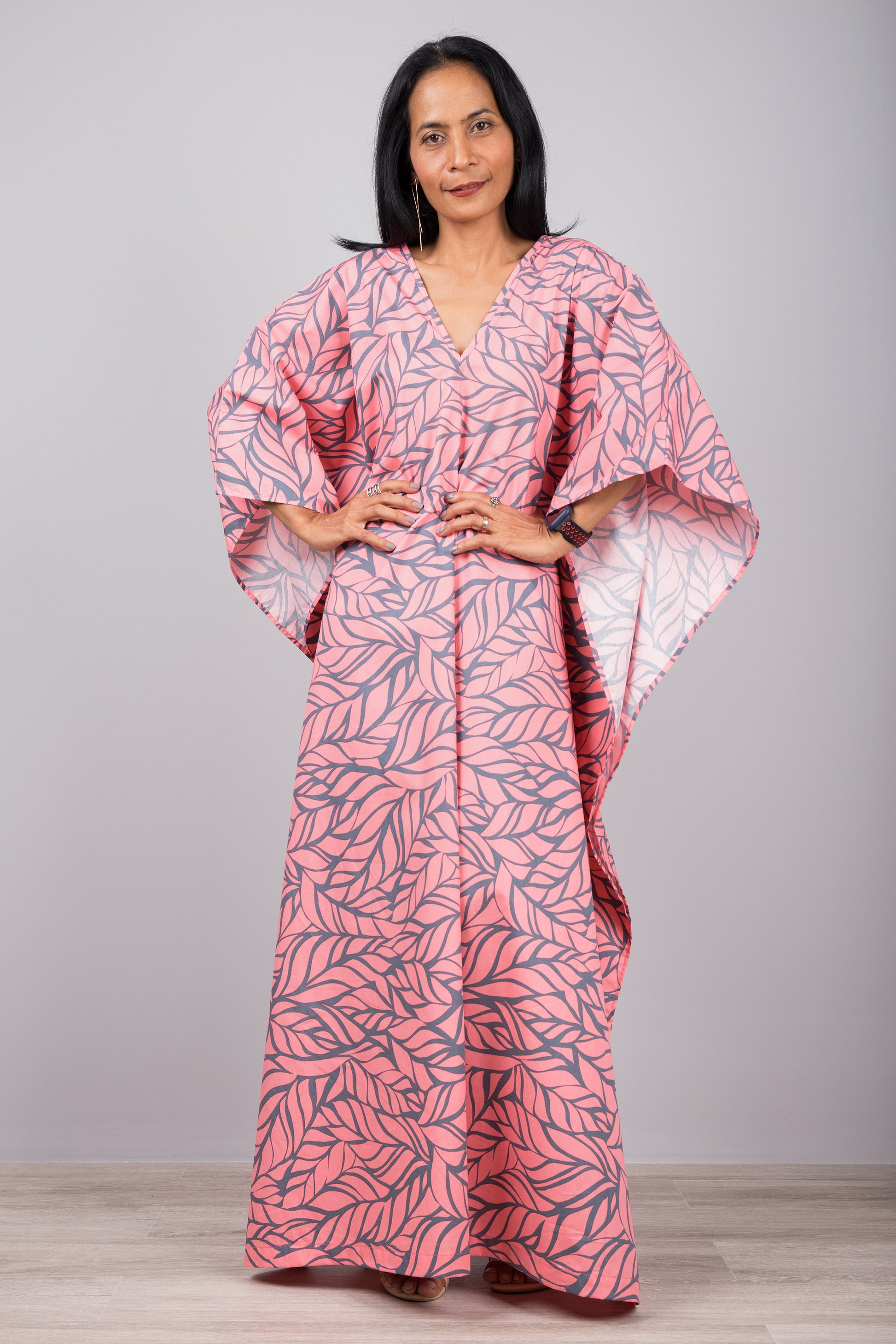 Woman in a pink leaf-patterned kaftan dress with side split, standing against a gray wall with a wooden floor, accessorized with a smartwatch and silver ring.

