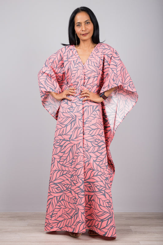 Woman in a pink leaf-patterned kaftan dress with side split, standing against a gray wall with a wooden floor, accessorized with a smartwatch and silver ring.

