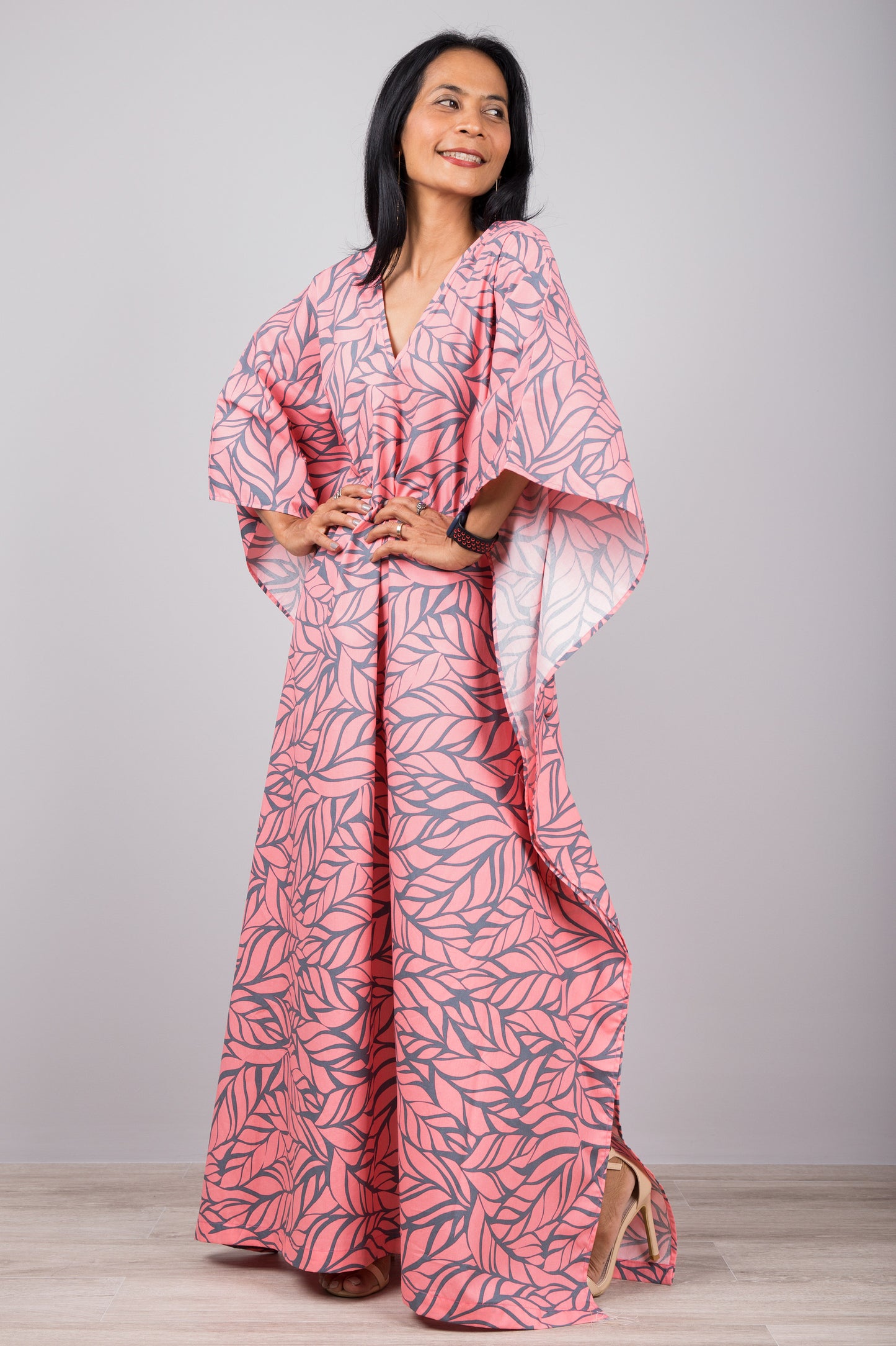 Woman in a soft pink kaftan dress with gray leaf print and high side split, posed against a gray wall with a wooden floor, wearing a smartwatch and red bracelet.

