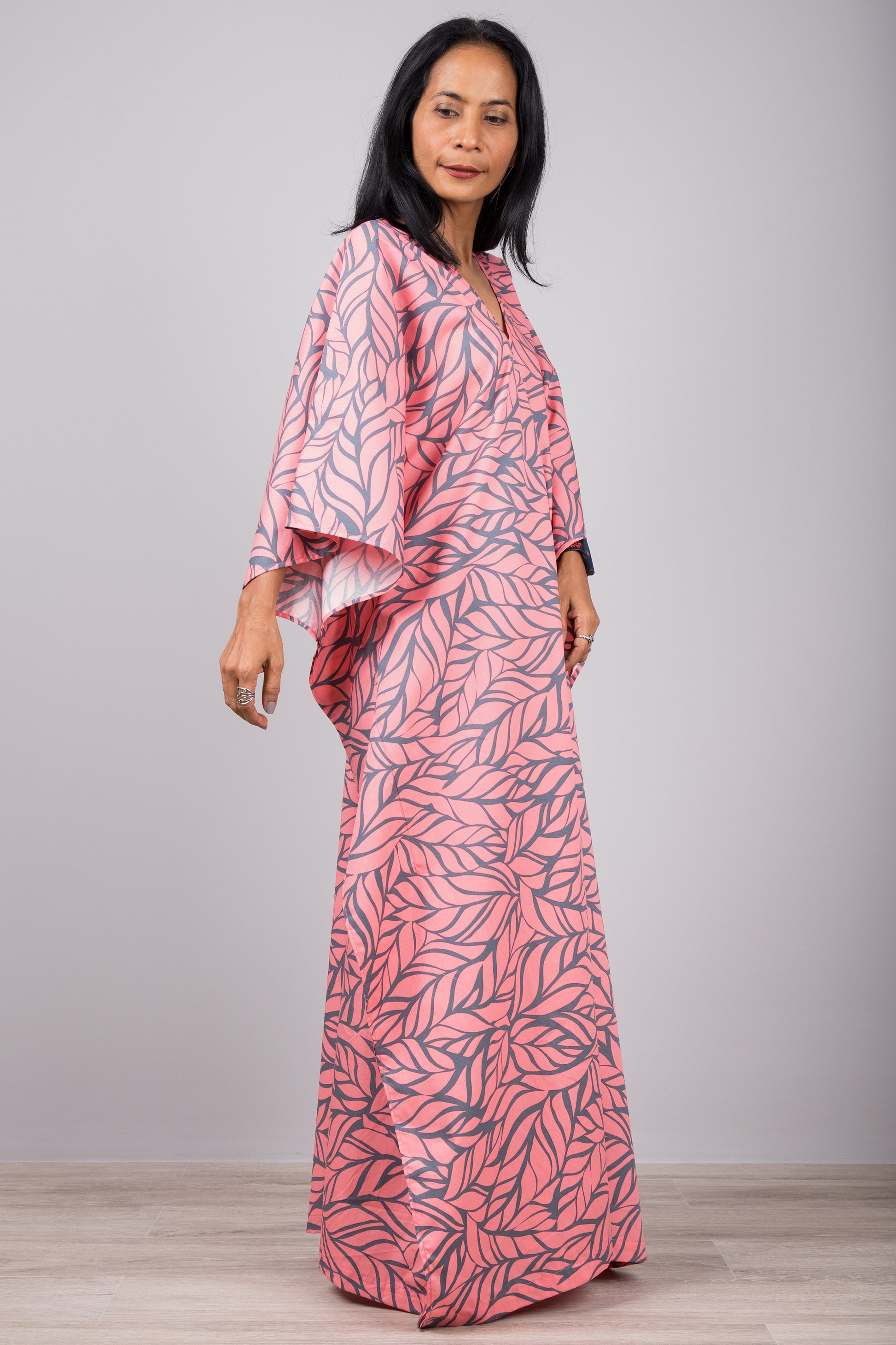 Woman in a boho-chic pink kaftan dress with tropical leaf design and side split, standing against a gray wall with a wooden floor, accessorized with a smartwatch and earrings.

