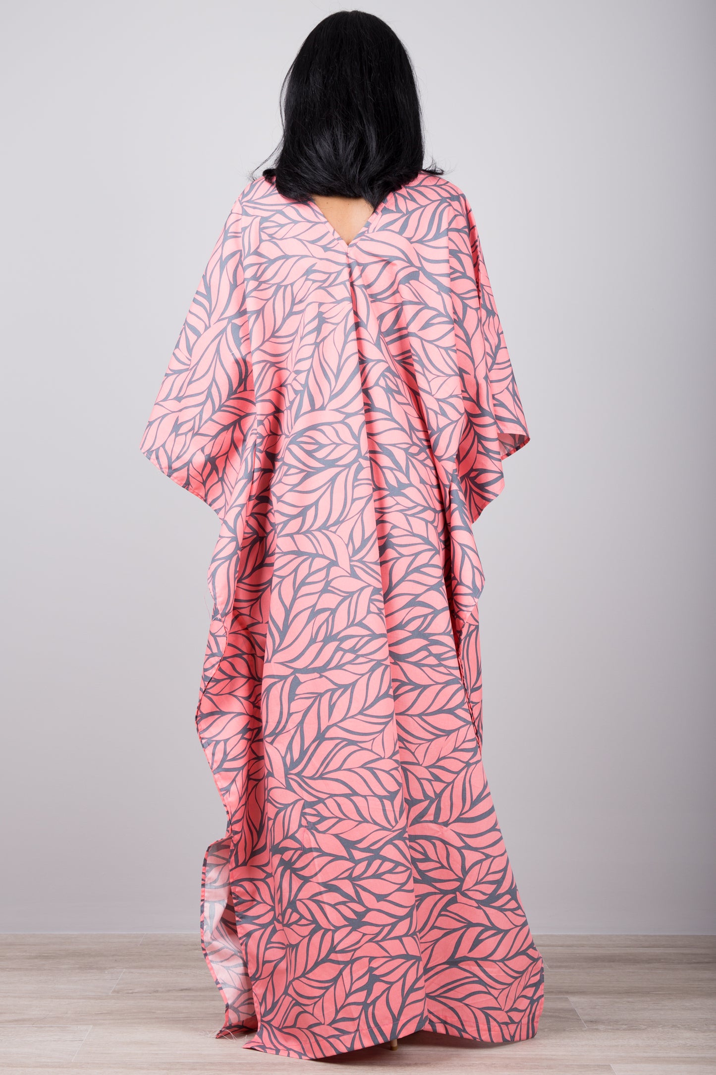 Woman in a lightweight pink kaftan dress with gray leaf pattern and side split, set against a gray wall with a wooden floor, wearing a smartwatch and silver cuff

