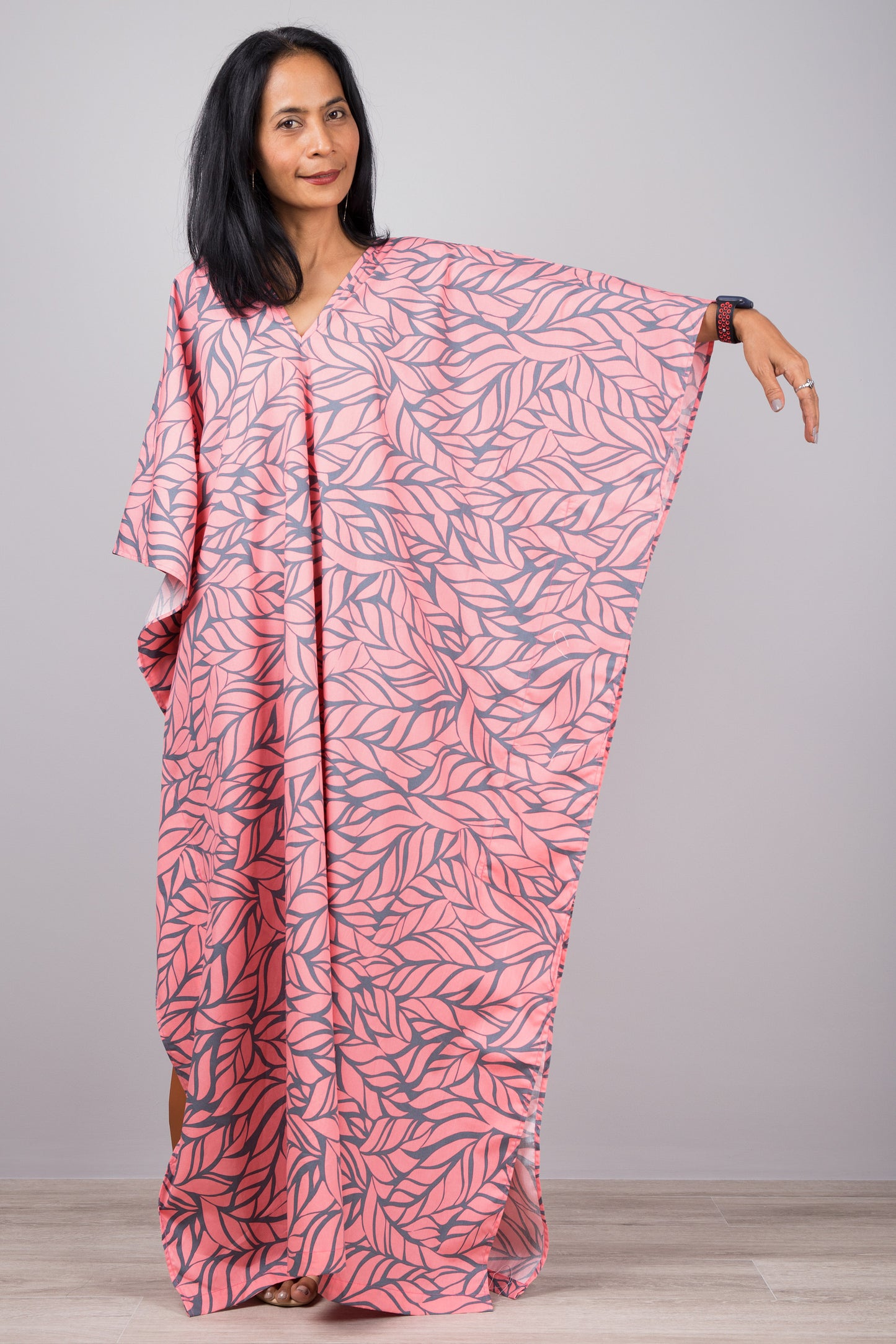Woman in a flowy pink kaftan dress with gray leaf print and side split, standing against a gray wall with a wooden floor, accessorized with a smartwatch and minimalist jewelry.

