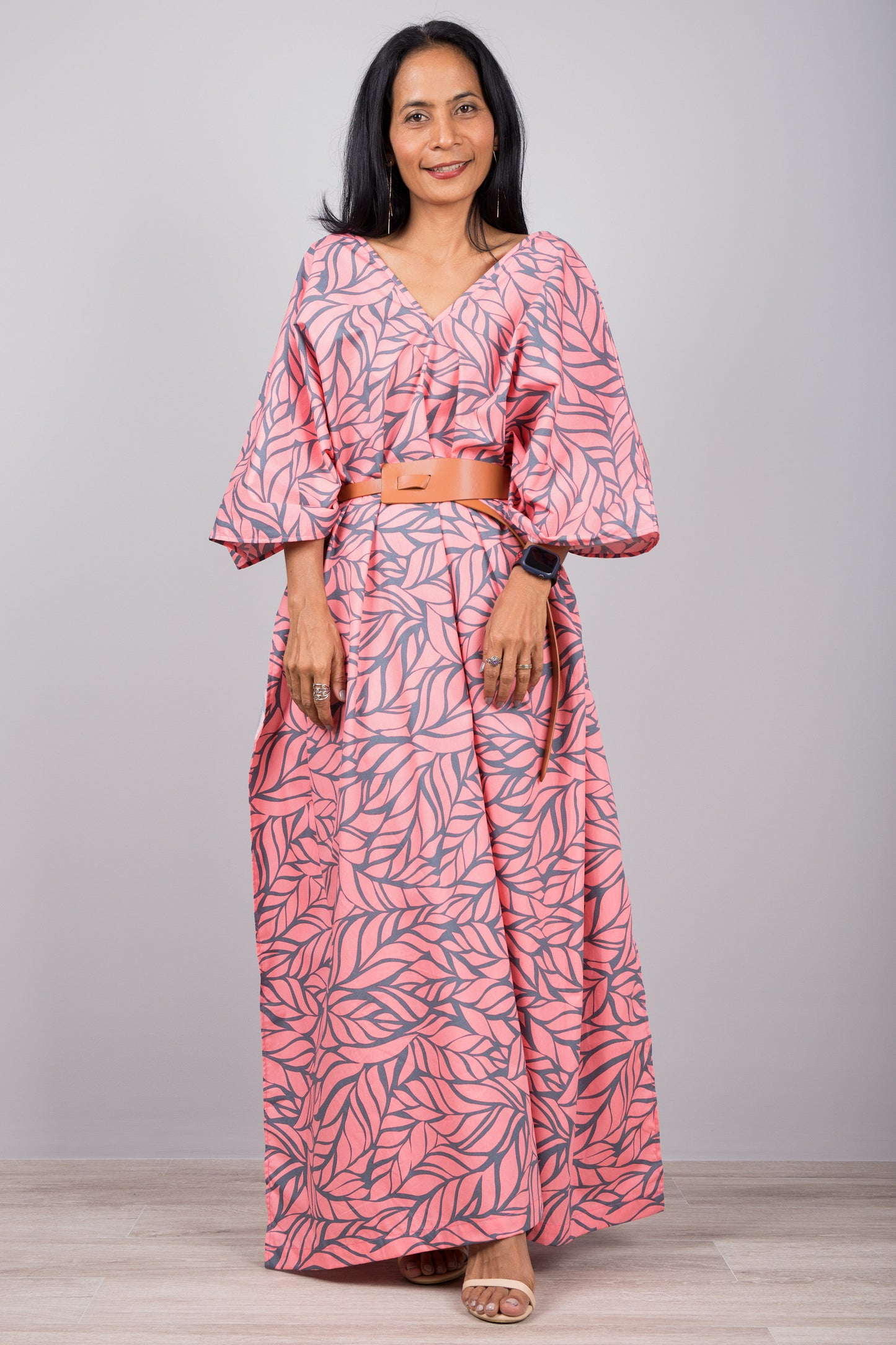 Woman in a one-size pink kaftan dress with tropical leaf pattern and side split, posed against a gray wall with a wooden floor, wearing a smartwatch and strappy sandals.

