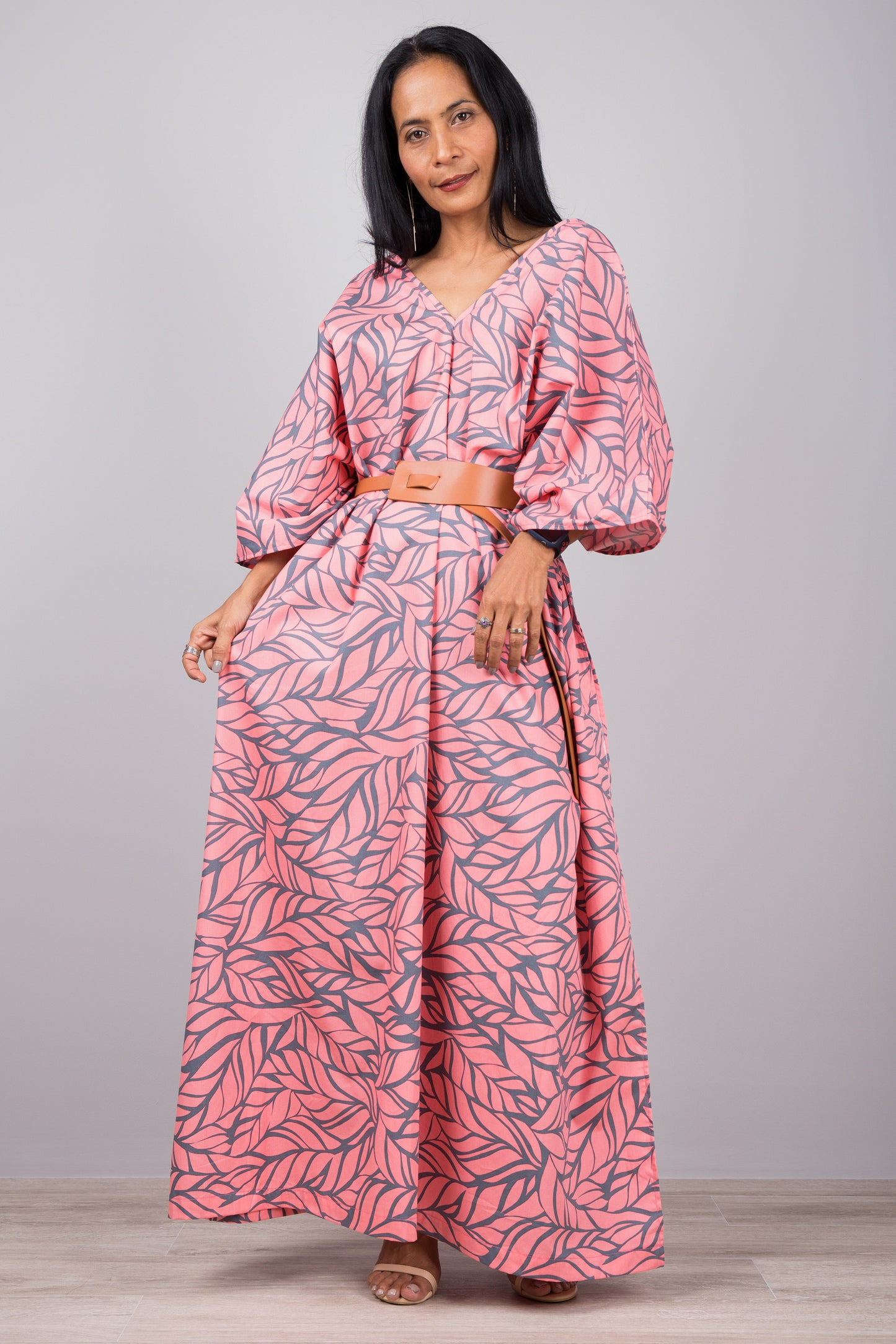 Woman in an elegant pink kaftan dress with gray leaf design and high side split, standing against a gray wall with a wooden floor, accessorized with a smartwatch and ring.

