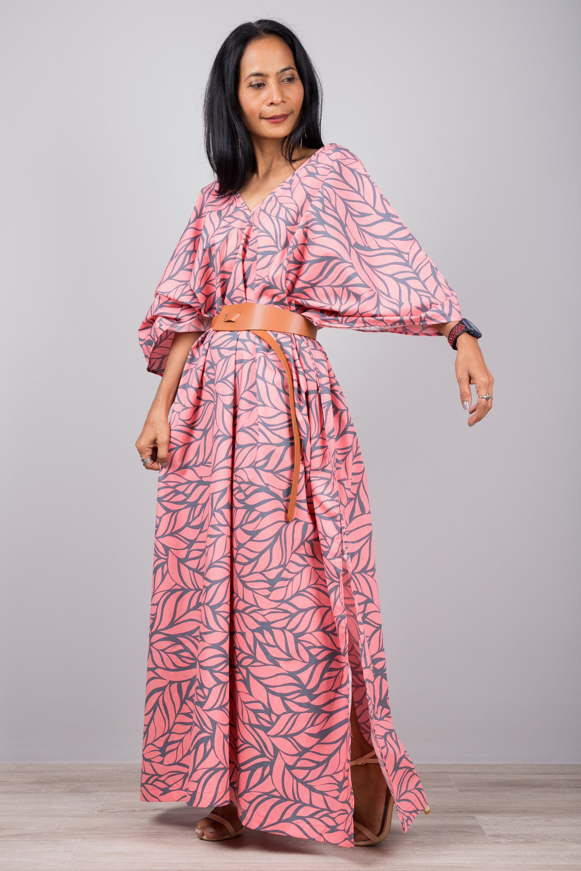 Woman in a summer-ready pink kaftan dress with gray leaf print and side split, set against a gray wall with a wooden floor, wearing a smartwatch and silver earrings.

