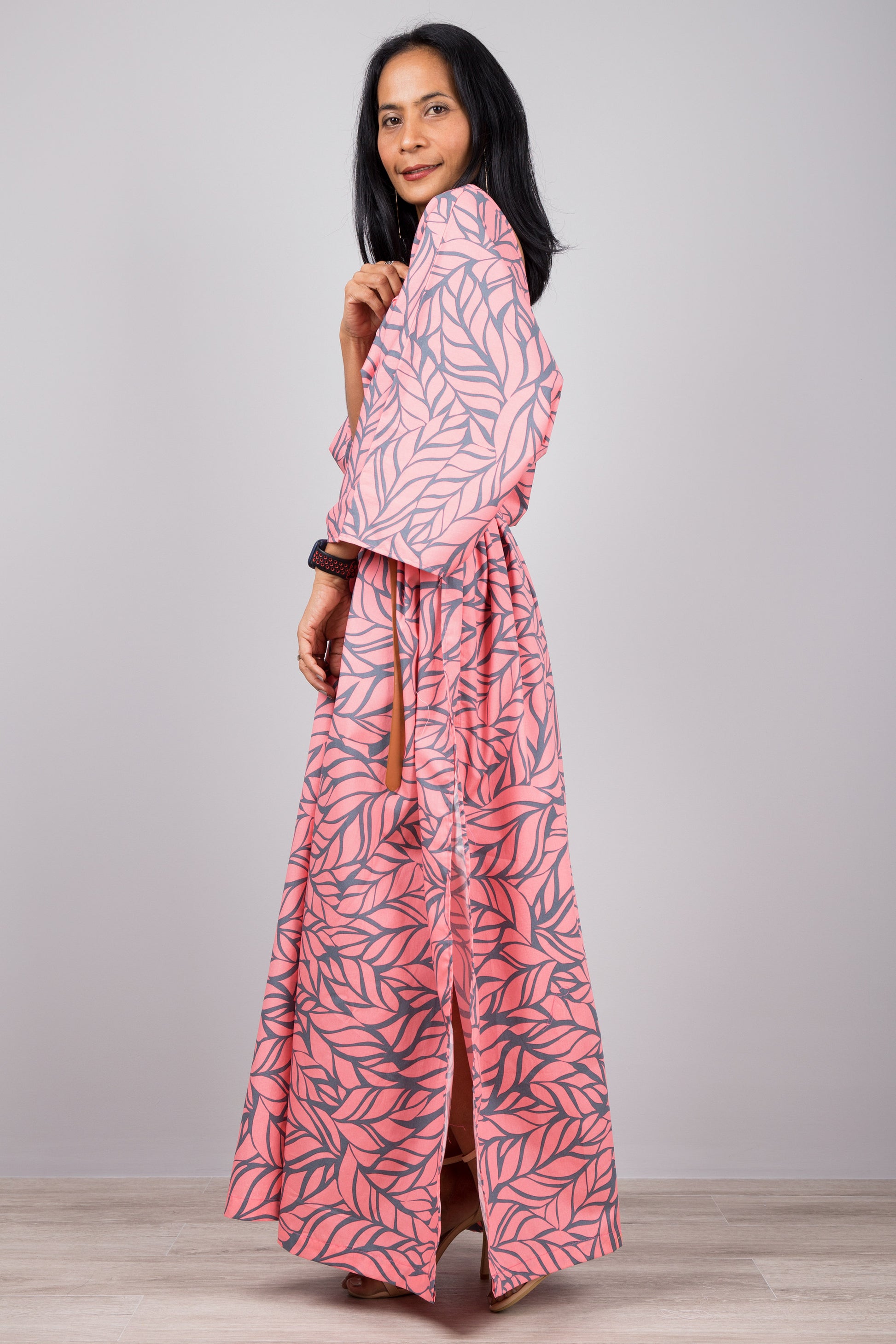 Woman in a versatile pink kaftan dress with gray leaf pattern and side split, standing against a gray wall with a wooden floor, accessorized with a smartwatch and bracelet.

