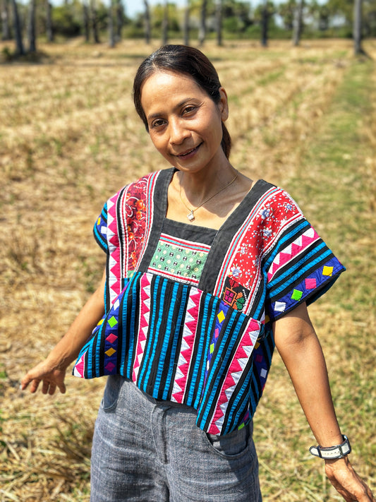 A woman wearing a multicolor tribal crop top in black, blue, magenta, and red, standing in a dry field with trees, featuring wide sleeves and geometric patterns, paired with gray pants and a white smartwatch