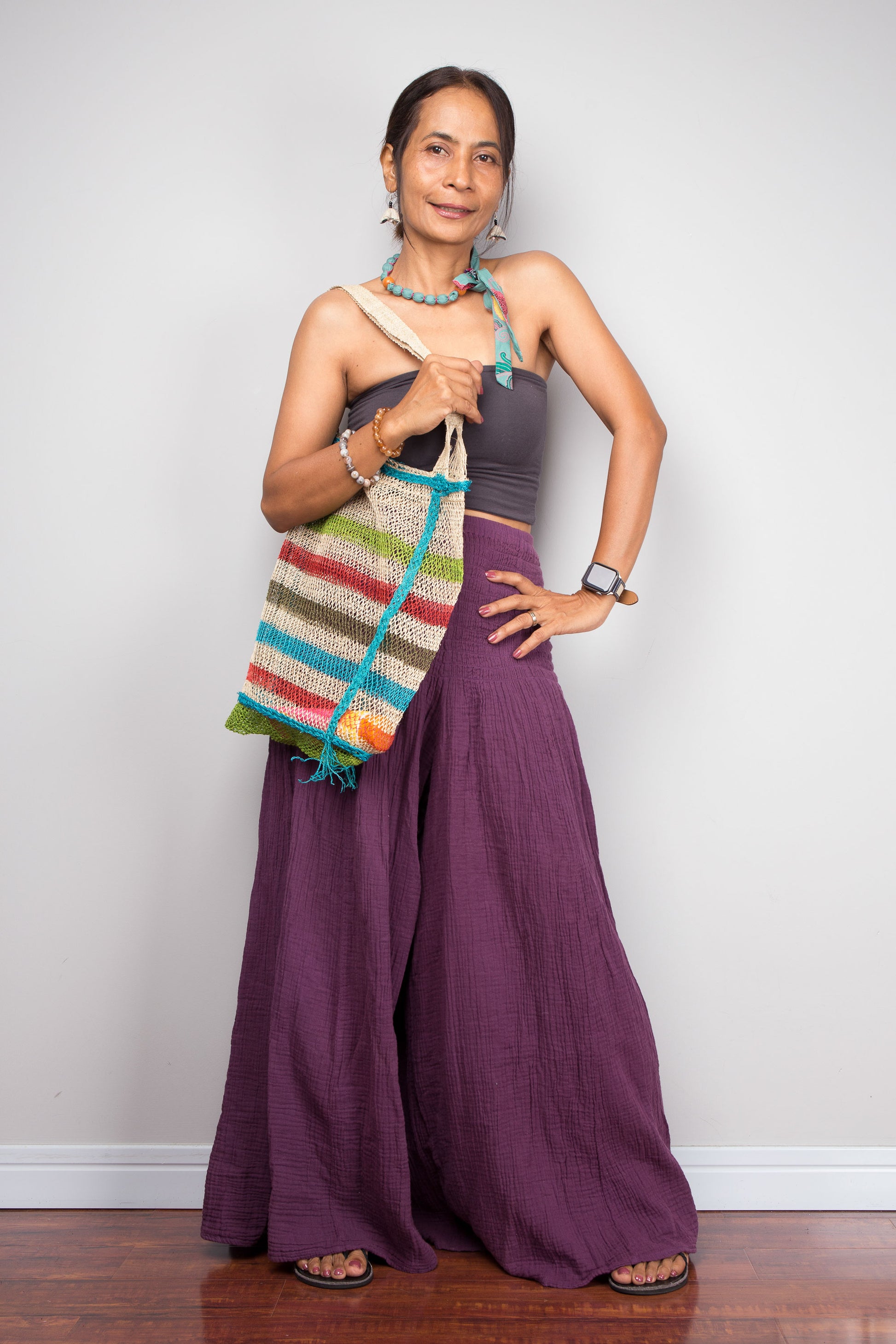 Woman in purple cotton pants holding a colorful striped bag against a white wall.