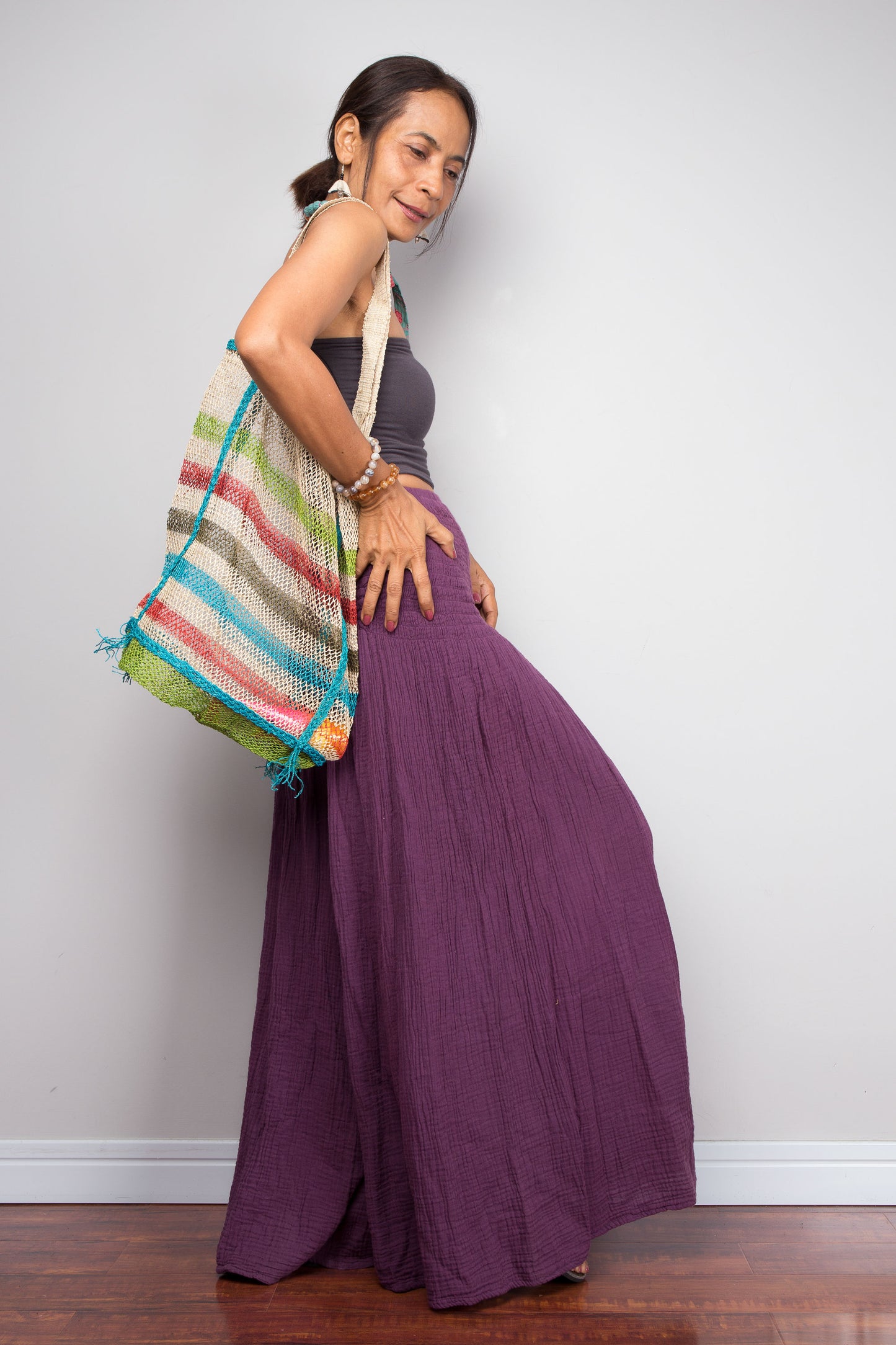 Woman wearing cotton purple pants ,holding a colorful striped bag against a plain background