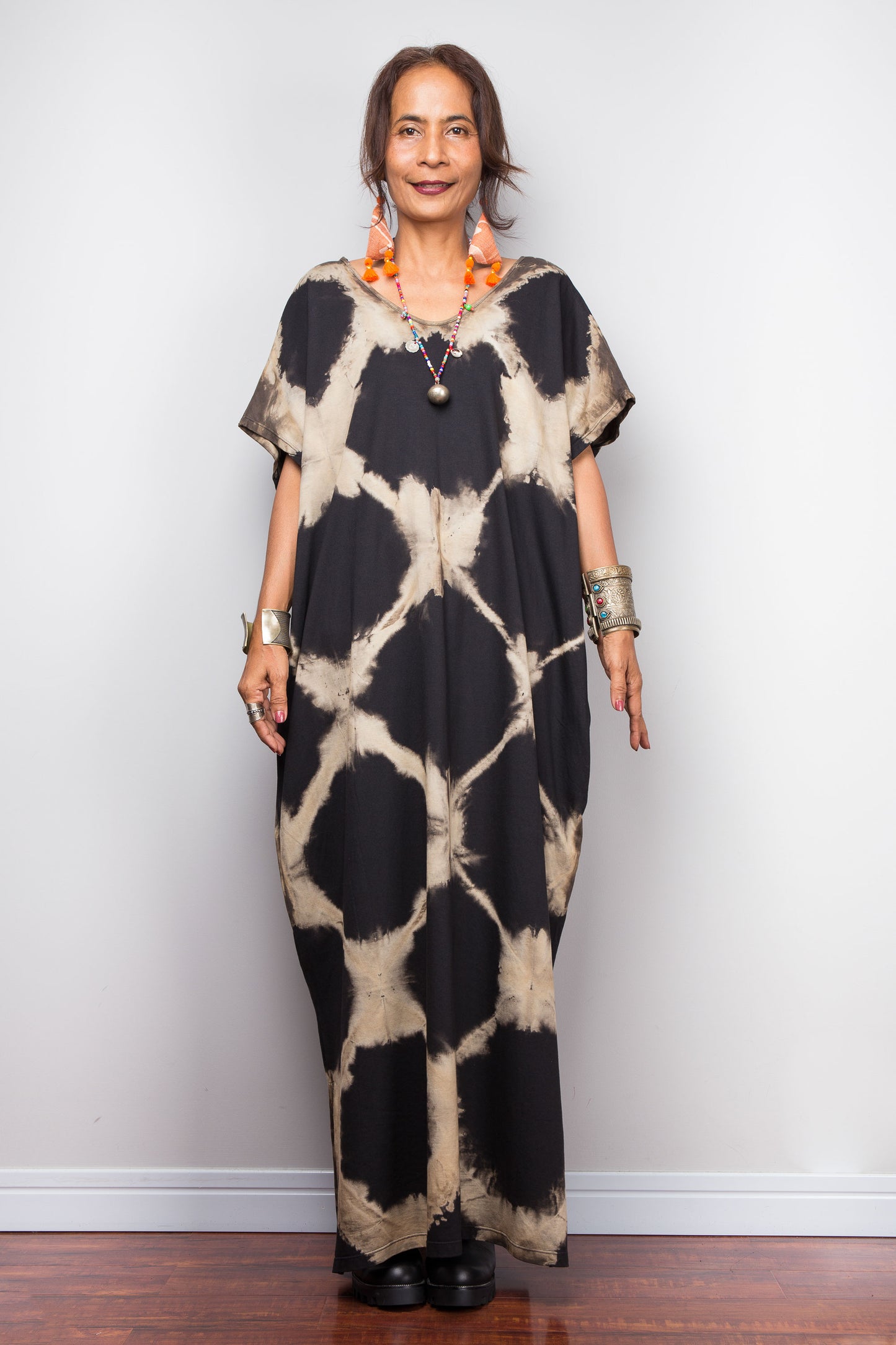 Woman wearing a black and beige patterned tie dye kaftan dress against a white wall.