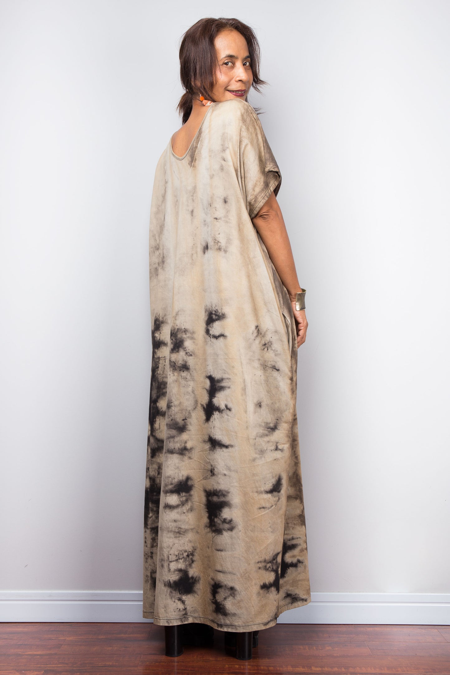 Full back view of a model in an authentic Nuichan brand reverse tie-dye kaftan maxi dress, featuring a soft sand and beige canvas with dark abstract watercolor bleeding patterns and a V-neckline.