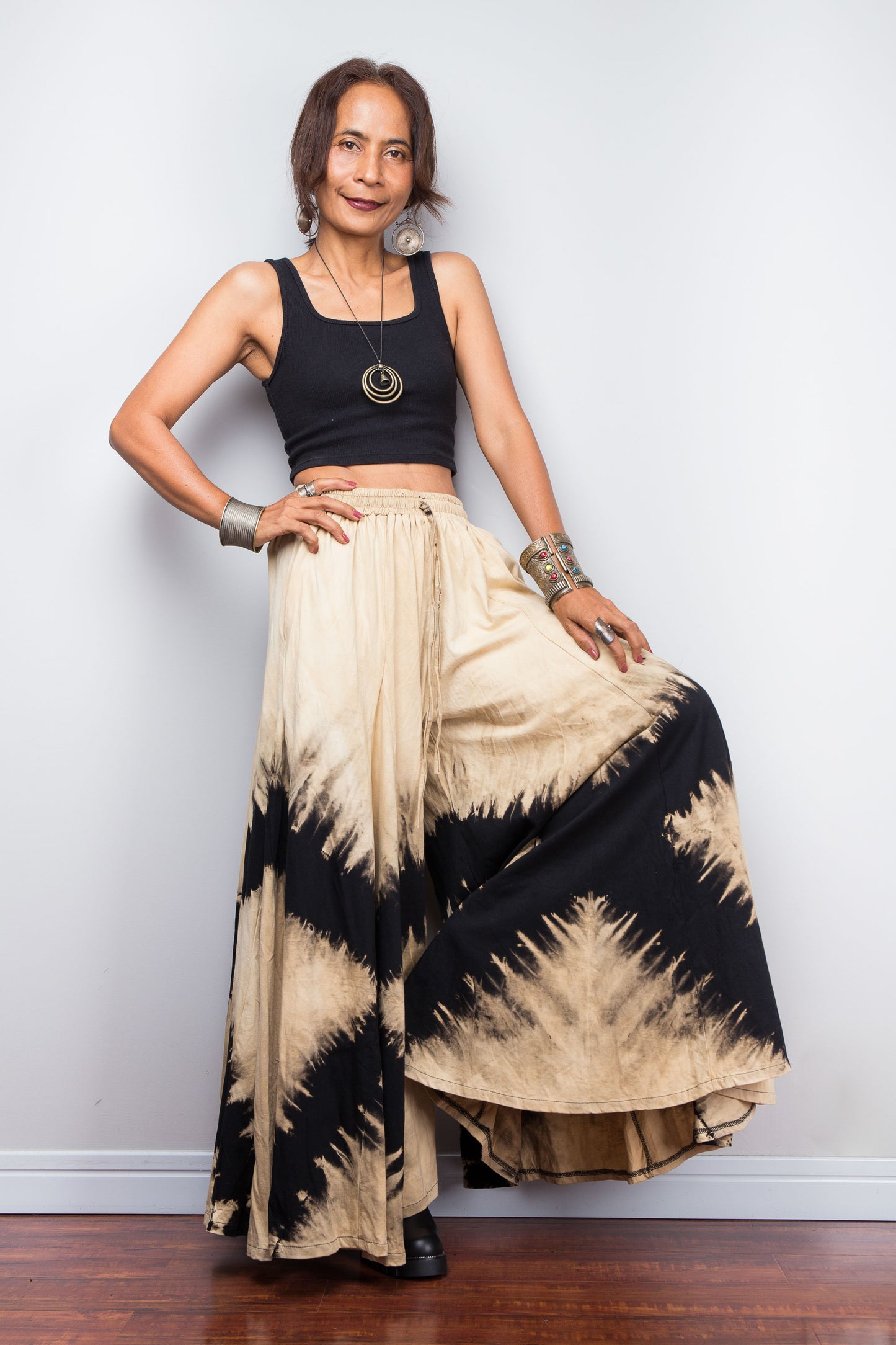 Petite Woman wearing a black and beige reverse tie dye pair of wide leg pants against a white wall.