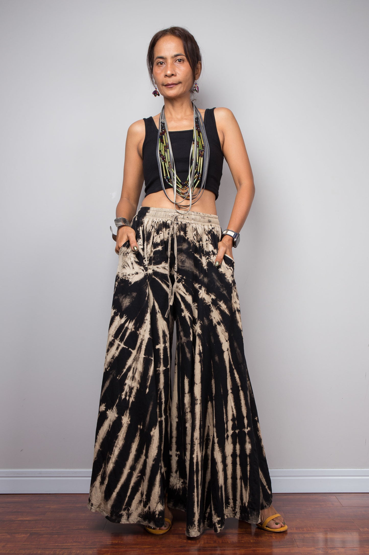 Front side view of a petite model wearing the Nuichan black and sand reverse tie dye wide leg palazzo pants in a vertical stripe pattern, styled high-waisted with a black crop top, showcasing the dramatic ultra-wide leg silhouette, the pockets, elongating vertical bleached stripes, and warm sand smocked elastic waistband with adjustable drawstring