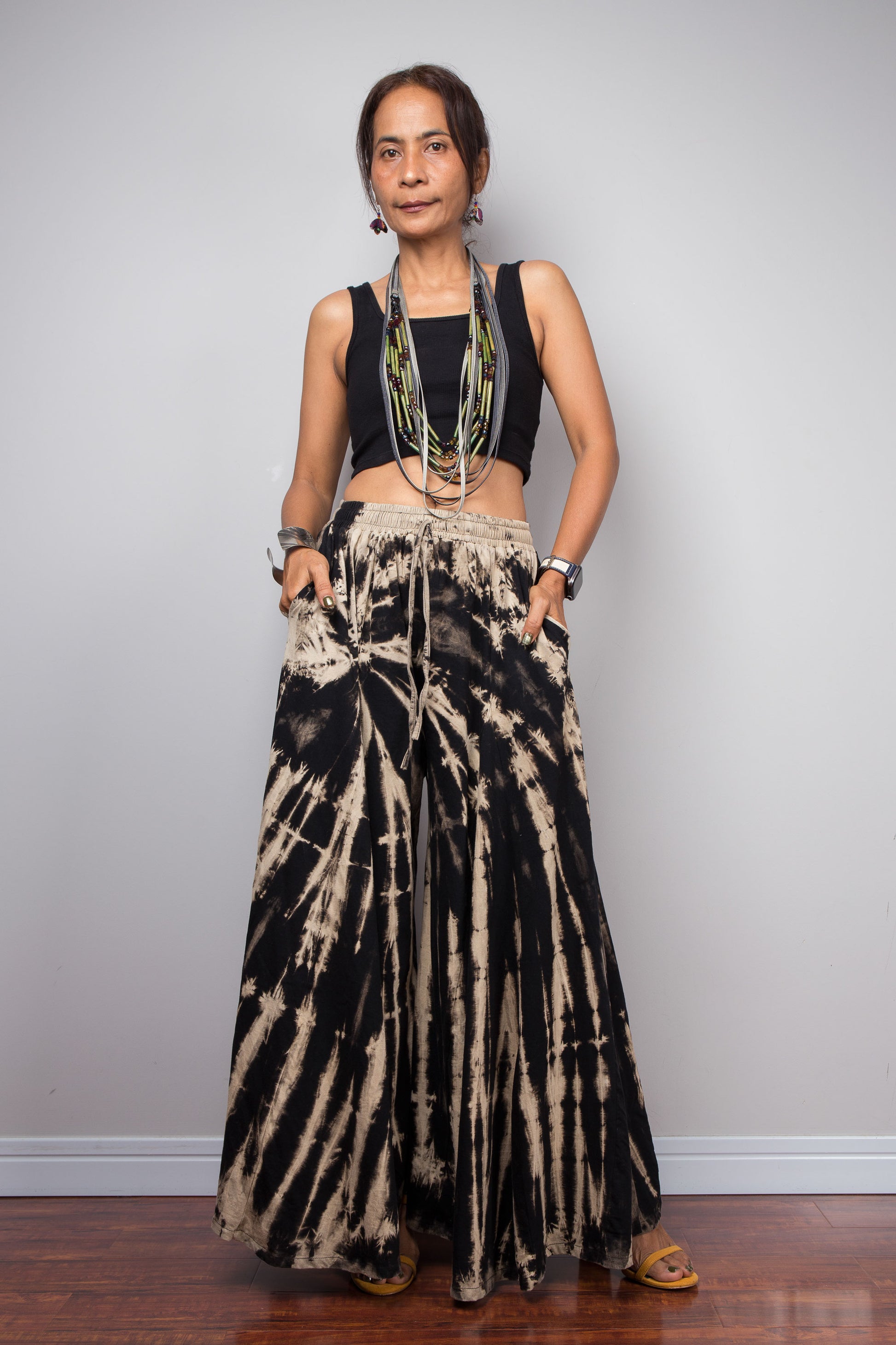 Front side view of a petite model wearing the Nuichan black and sand reverse tie dye wide leg palazzo pants in a vertical stripe pattern, styled high-waisted with a black crop top, showcasing the dramatic ultra-wide leg silhouette, the pockets, elongating vertical bleached stripes, and warm sand smocked elastic waistband with adjustable drawstring