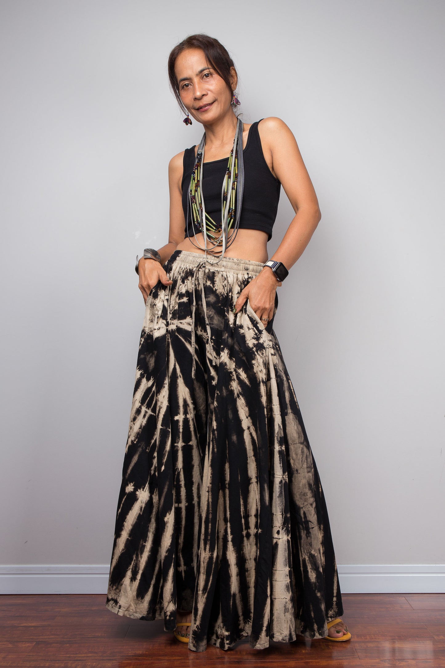 Front view of a petite model wearing the Nuichan black and sand reverse tie dye wide leg palazzo pants in a vertical stripe pattern, styled high-waisted with a black crop top, showcasing the dramatic ultra-wide leg silhouette, the inseam pockets, elongating vertical bleached stripes, and warm sand smocked elastic waistband with adjustable drawstring