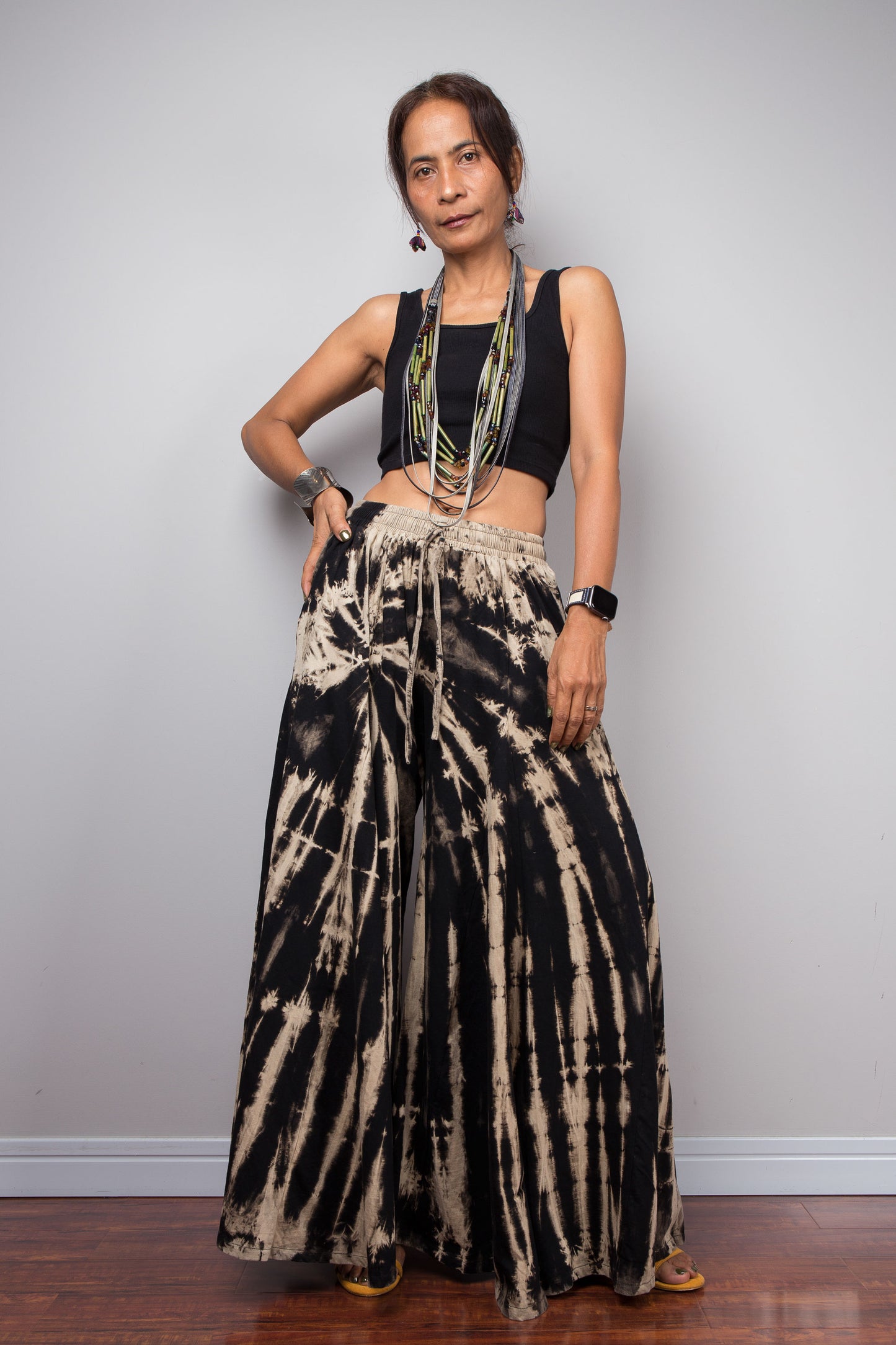 Front view of a petite model in the Nuichan black and sand stripe reverse tie dye palazzo pants with hand on the hip demonstrating the immense skirt-like volume and fluid drape of the ultra-wide cotton legs