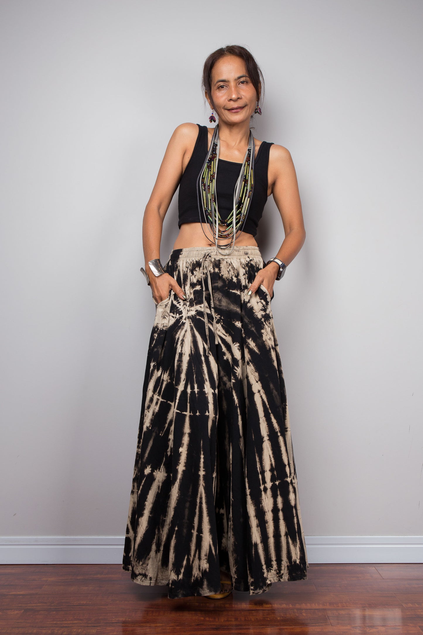 Front view of a petite female model wearing the Nuichan black and sand reverse tie dye wide leg palazzo pants in a vertical stripe pattern, styled high-waisted with a black crop top, showcasing the dramatic ultra-wide leg silhouette, elongating vertical bleached stripes, the inseam pockets and warm sand smocked elastic waistband with adjustable drawstring