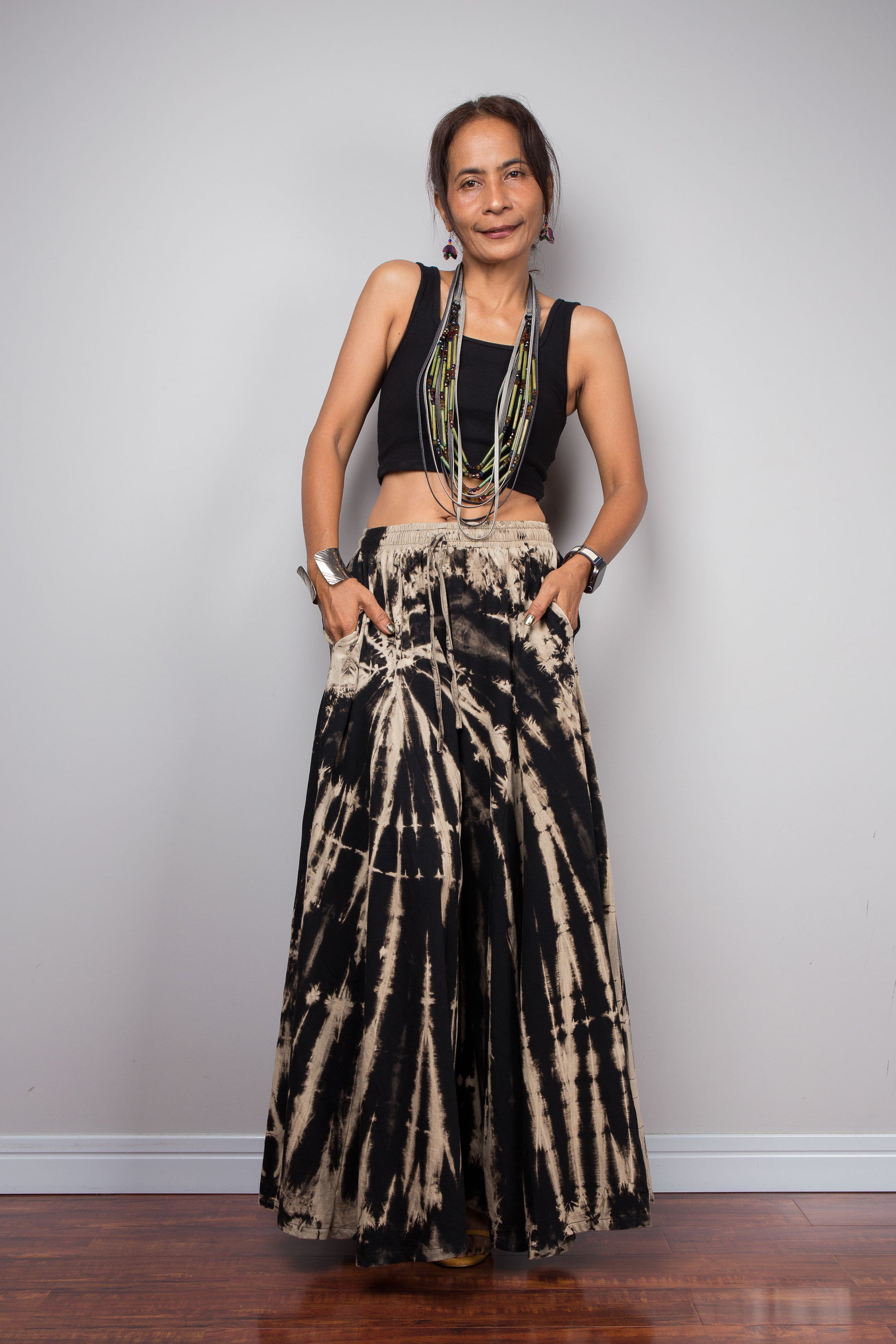 Front view of a petite female model wearing the Nuichan black and sand reverse tie dye wide leg palazzo pants in a vertical stripe pattern, styled high-waisted with a black crop top, showcasing the dramatic ultra-wide leg silhouette, elongating vertical bleached stripes, the inseam pockets and warm sand smocked elastic waistband with adjustable drawstring