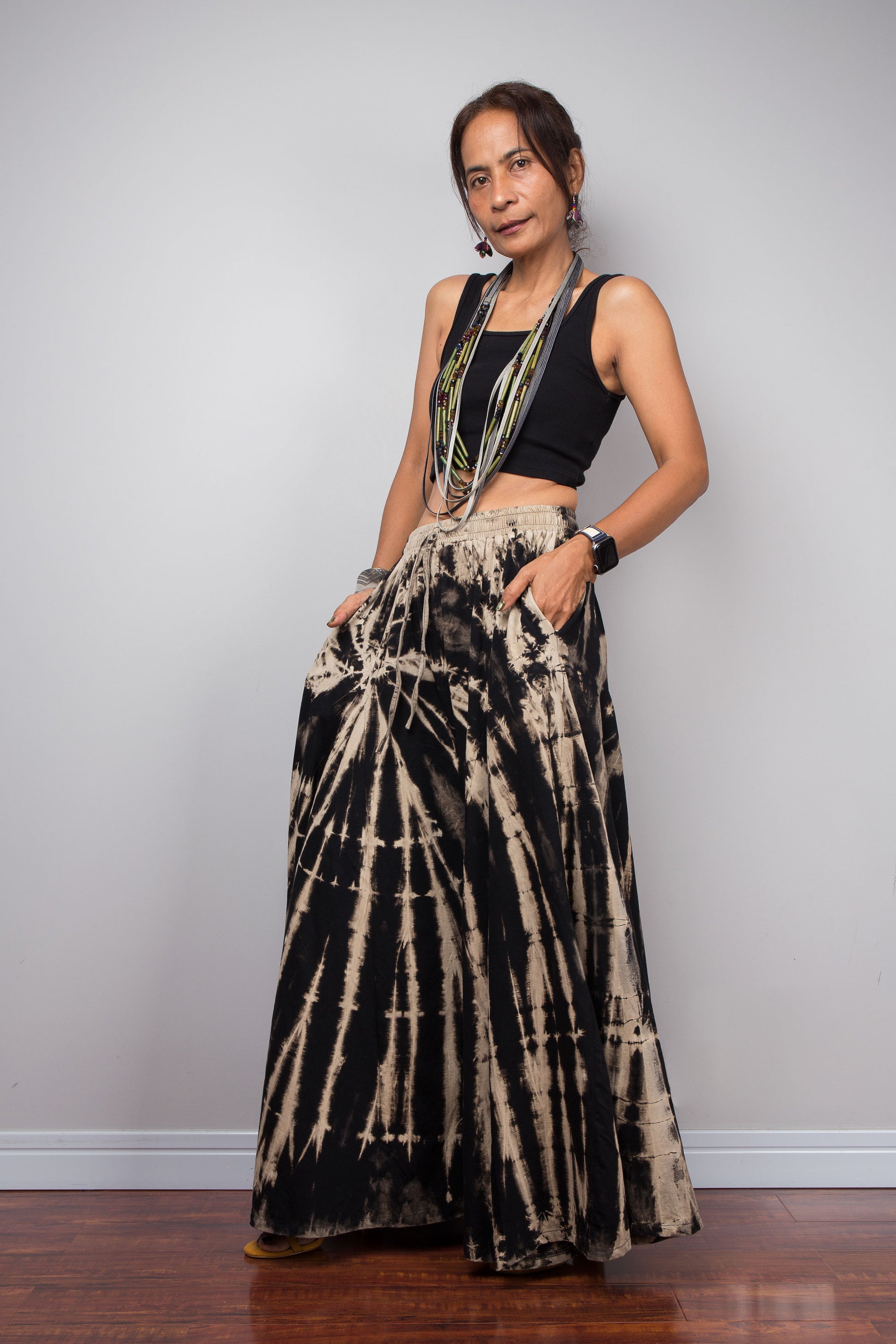 Three-quarter view of a model wearing the Nuichan black and sand reverse tie dye wide leg palazzo pants in a vertical shibori stripe pattern, styled high-waisted with a black crop top, showcasing the dramatic ultra-wide leg silhouette, elongating vertical bleached stripes, and warm sand smocked elastic waistband with adjustable drawstring